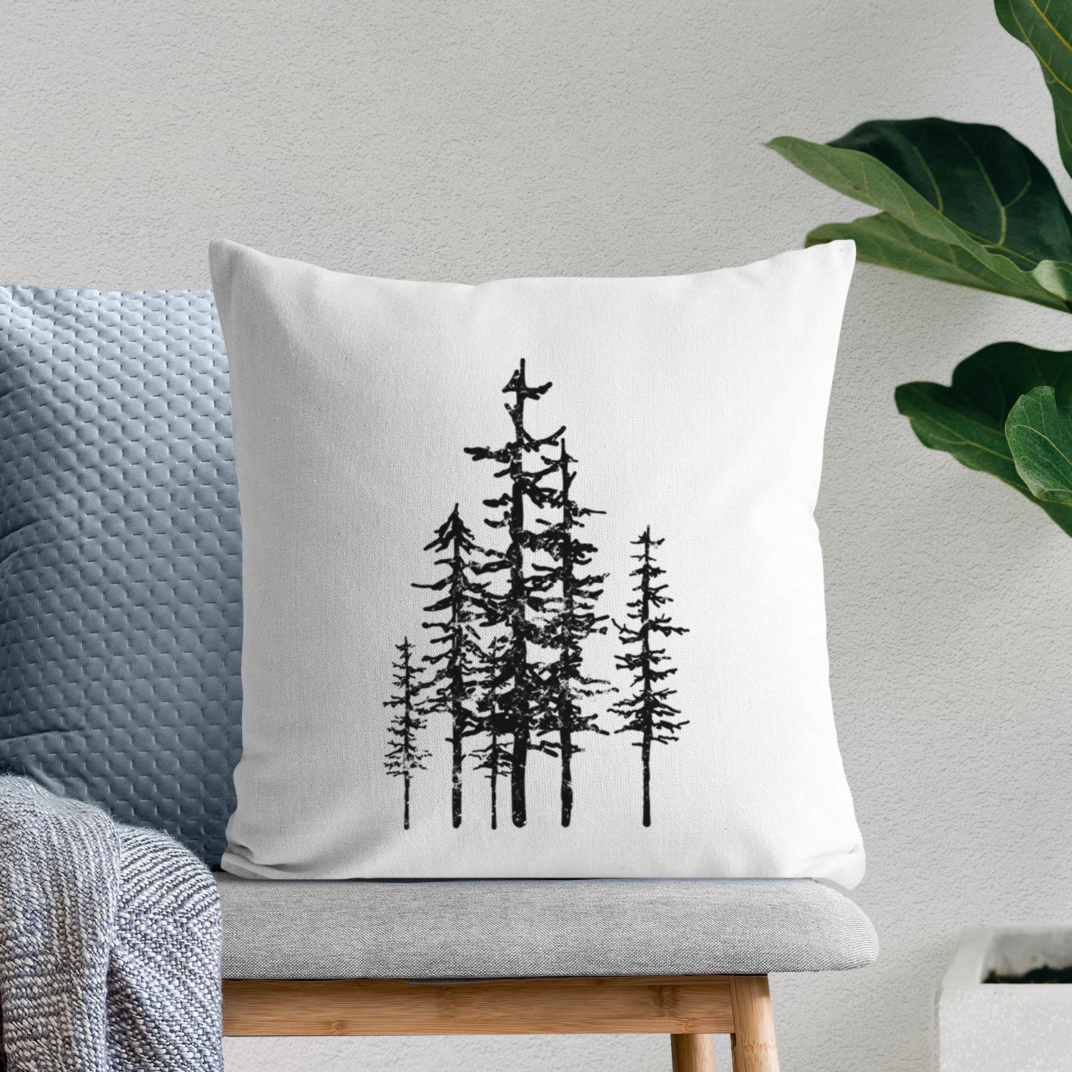 White pillow cover printed with a black ink illustration of a cluster of evergreen trees of varying heights, on a bench beside plants.