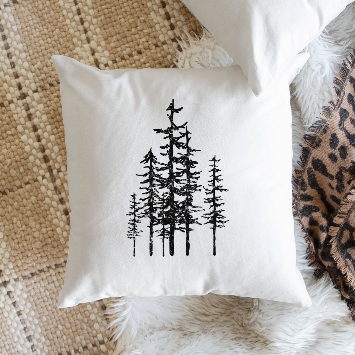 White pillow cover printed with a black ink illustration of a cluster of evergreen trees of varying heights, on a woven chair.