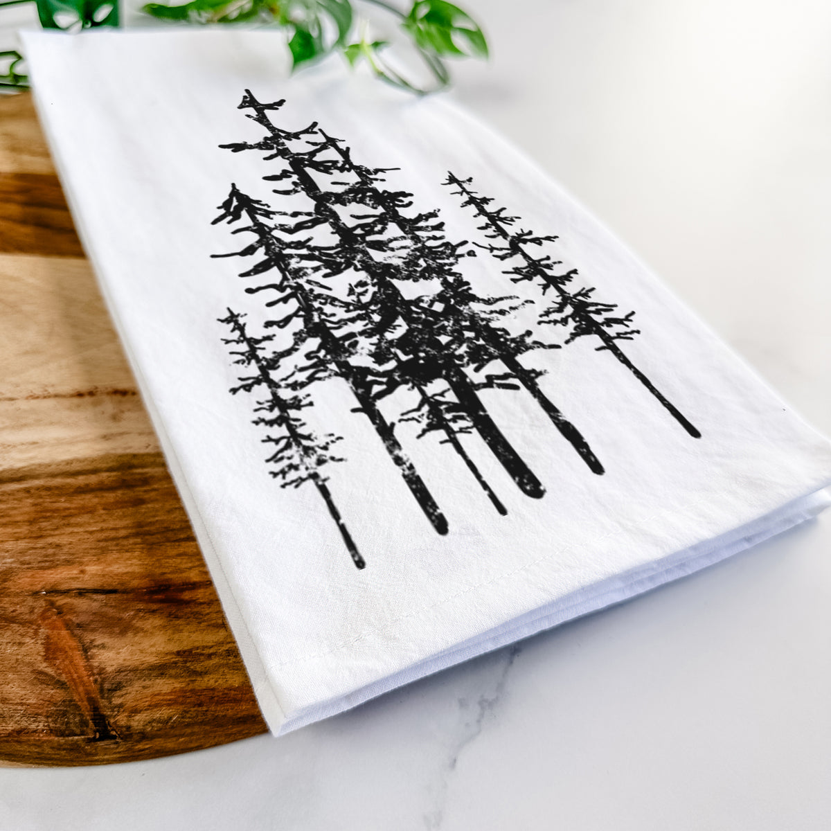 Close-up of a folded white tea towel on a wooden board, showing a black ink illustration of a cluster of evergreen trees of varying heights.