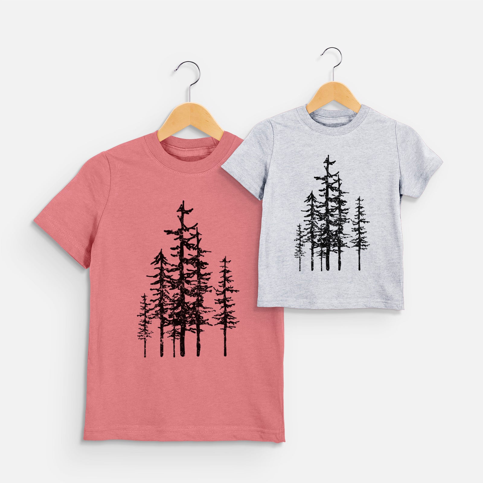 Two youth t-shirts in different colors on hangers, printed with a black ink illustration of a cluster of evergreen trees of varying heights.