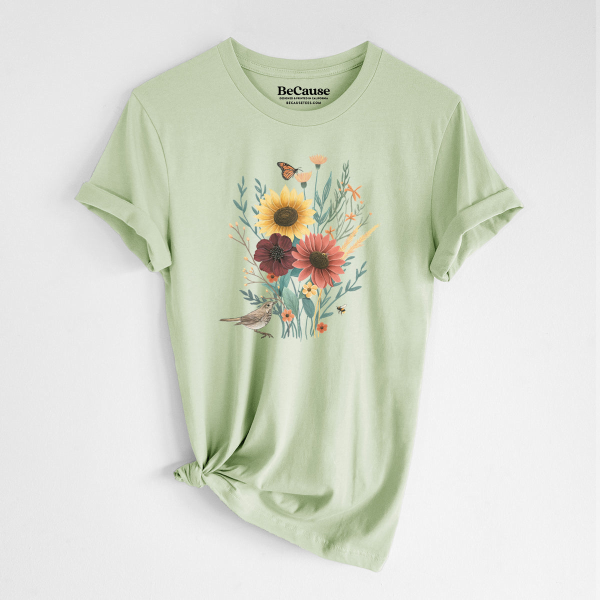 Spring green lightweight cotton t-shirt laid flat with the sleeves rolled, printed with a full-color bouquet of sunflowers, red flowers and wildflowers with butterflies and a small bird.