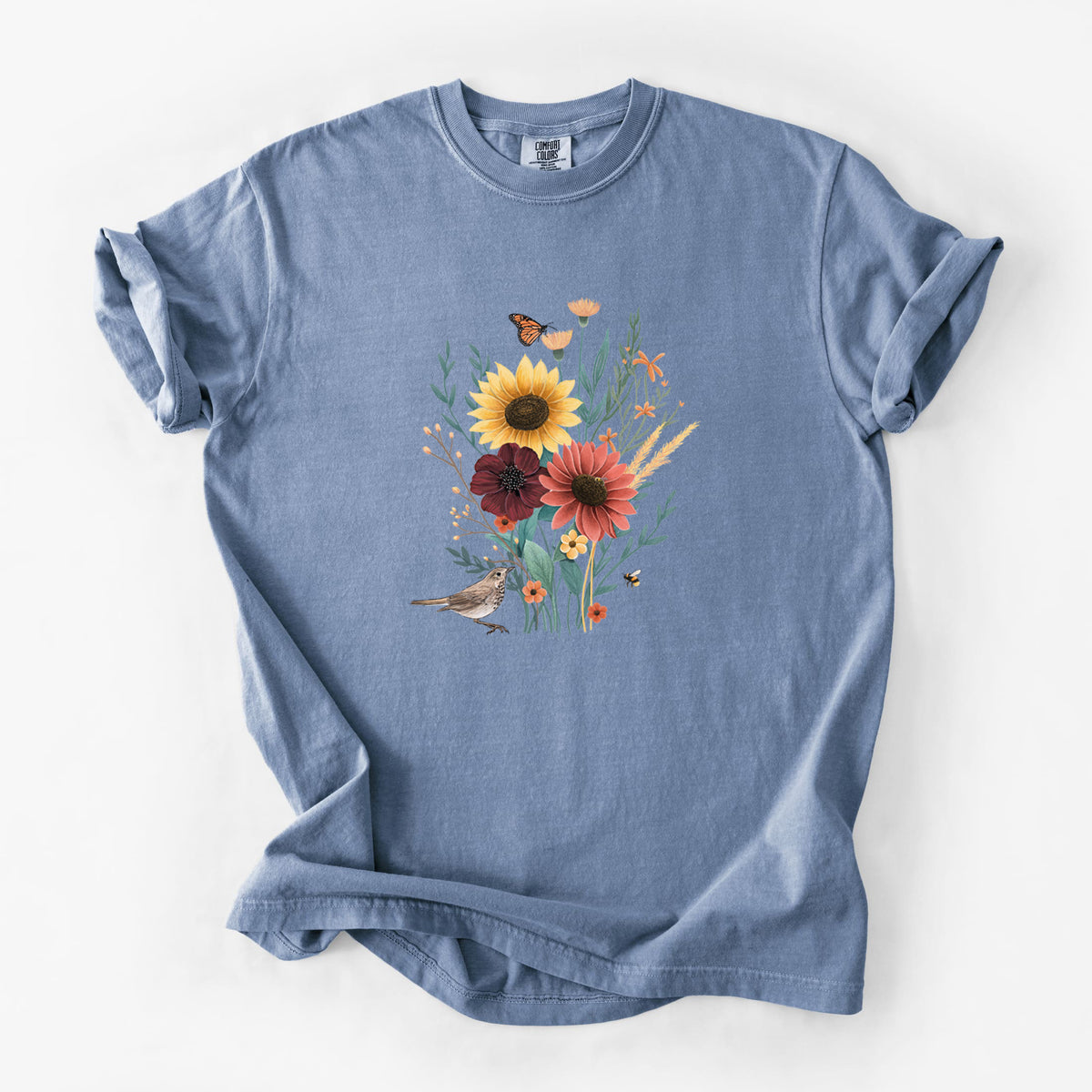 Blue jean Comfort Colors t-shirt laid flat, printed with a full-color bouquet of sunflowers, red flowers and wildflowers with butterflies and a small bird.