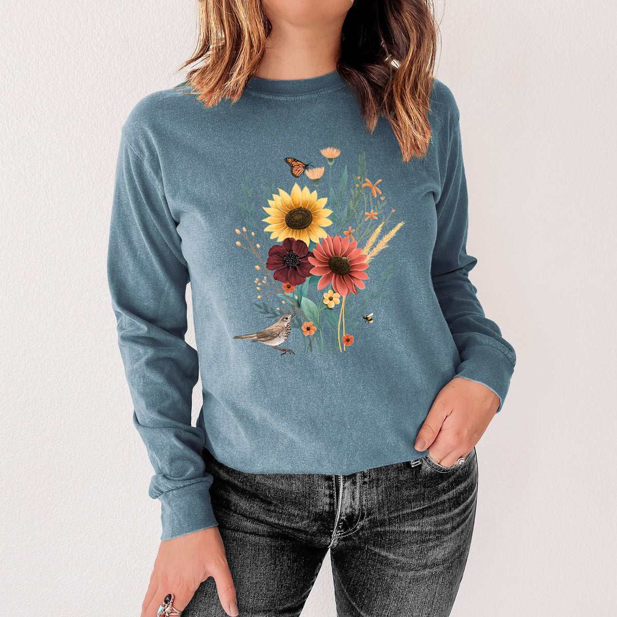 Steel blue Comfort Colors long sleeve shirt printed with a full-color bouquet of sunflowers, red flowers and wildflowers with butterflies and a small bird, shown in a lifestyle photo.
