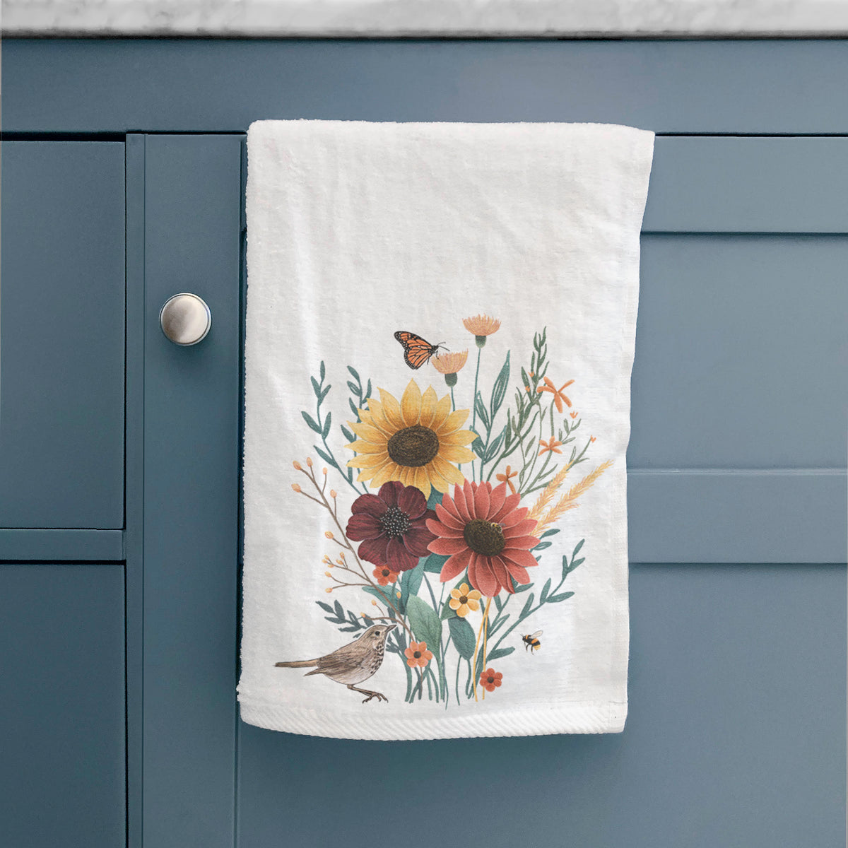 White hand towel printed with a full-color bouquet of sunflowers, red flowers and wildflowers with butterflies and a small bird, hanging on a cabinet door.