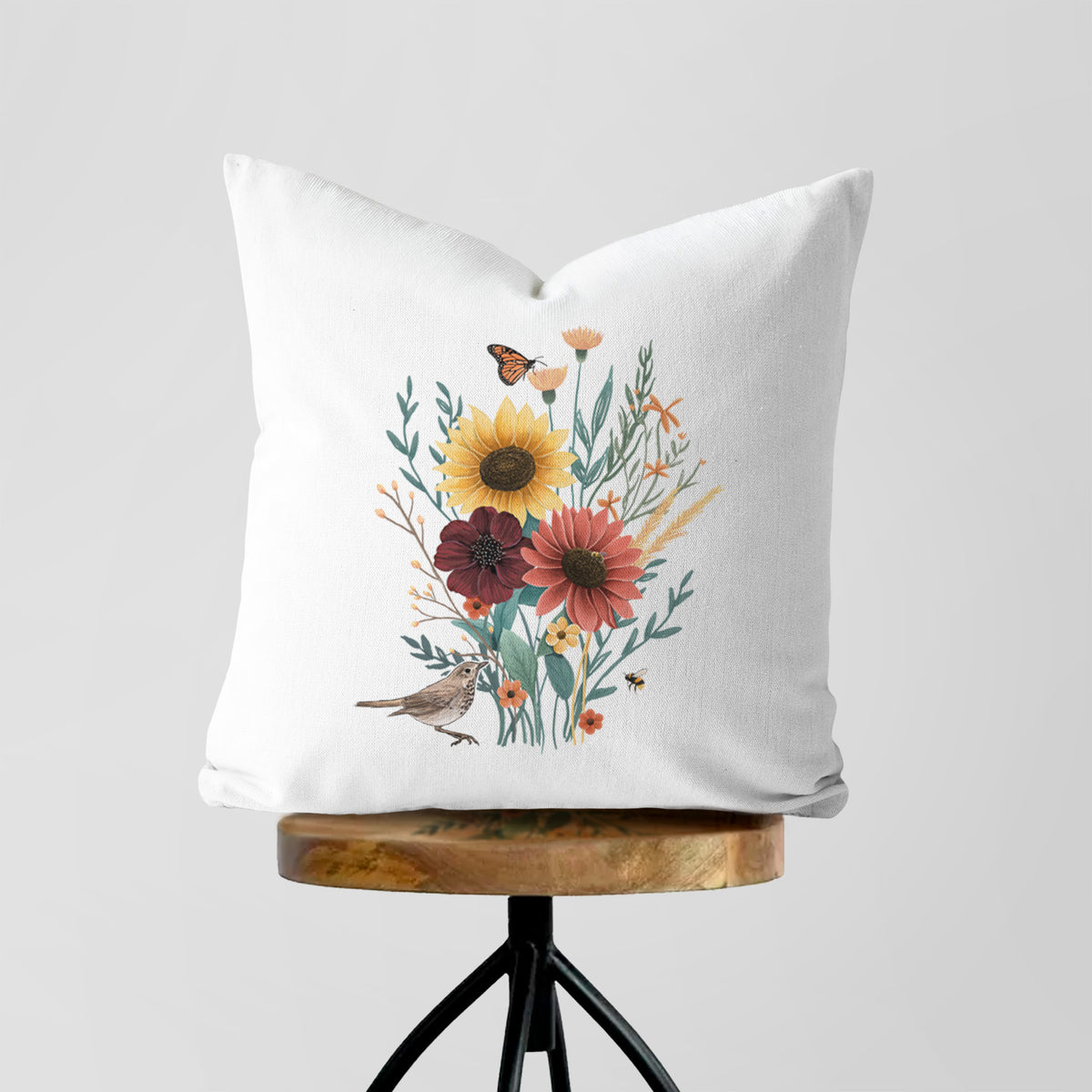 White pillow cover printed with a full-color bouquet of sunflowers, red flowers and wildflowers with butterflies and a small bird, on a wooden stool.