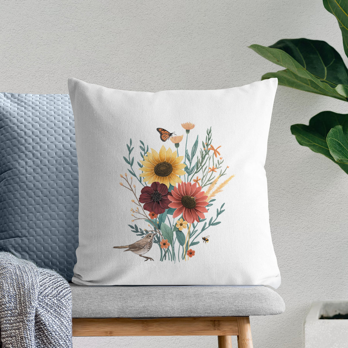 White pillow cover printed with a full-color bouquet of sunflowers, red flowers and wildflowers with butterflies and a small bird, on a bench beside plants.