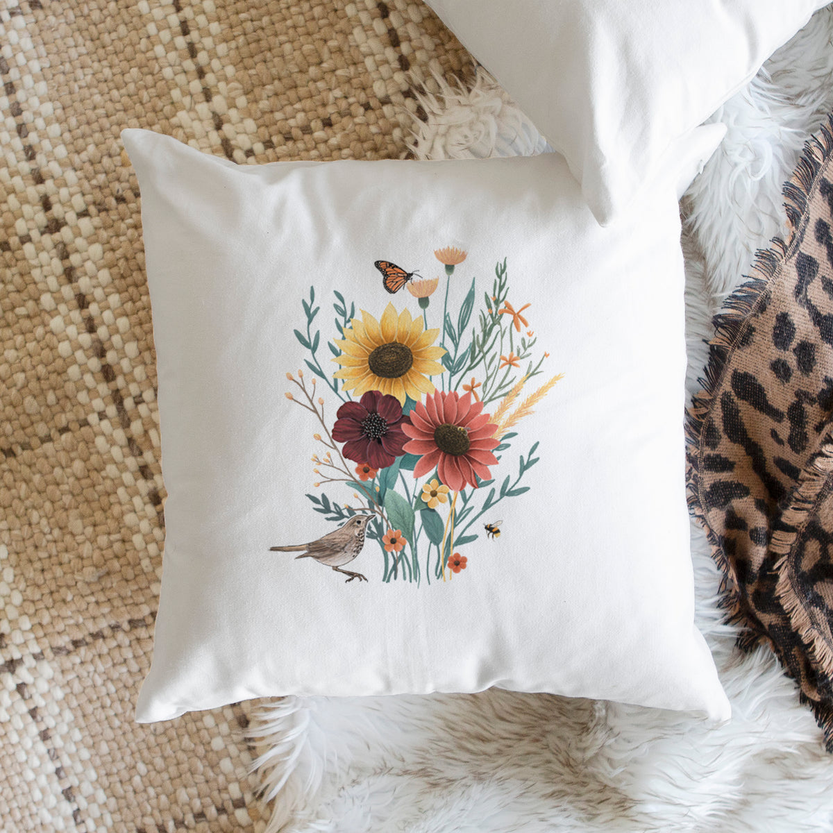 White pillow cover printed with a full-color bouquet of sunflowers, red flowers and wildflowers with butterflies and a small bird, on a woven chair.