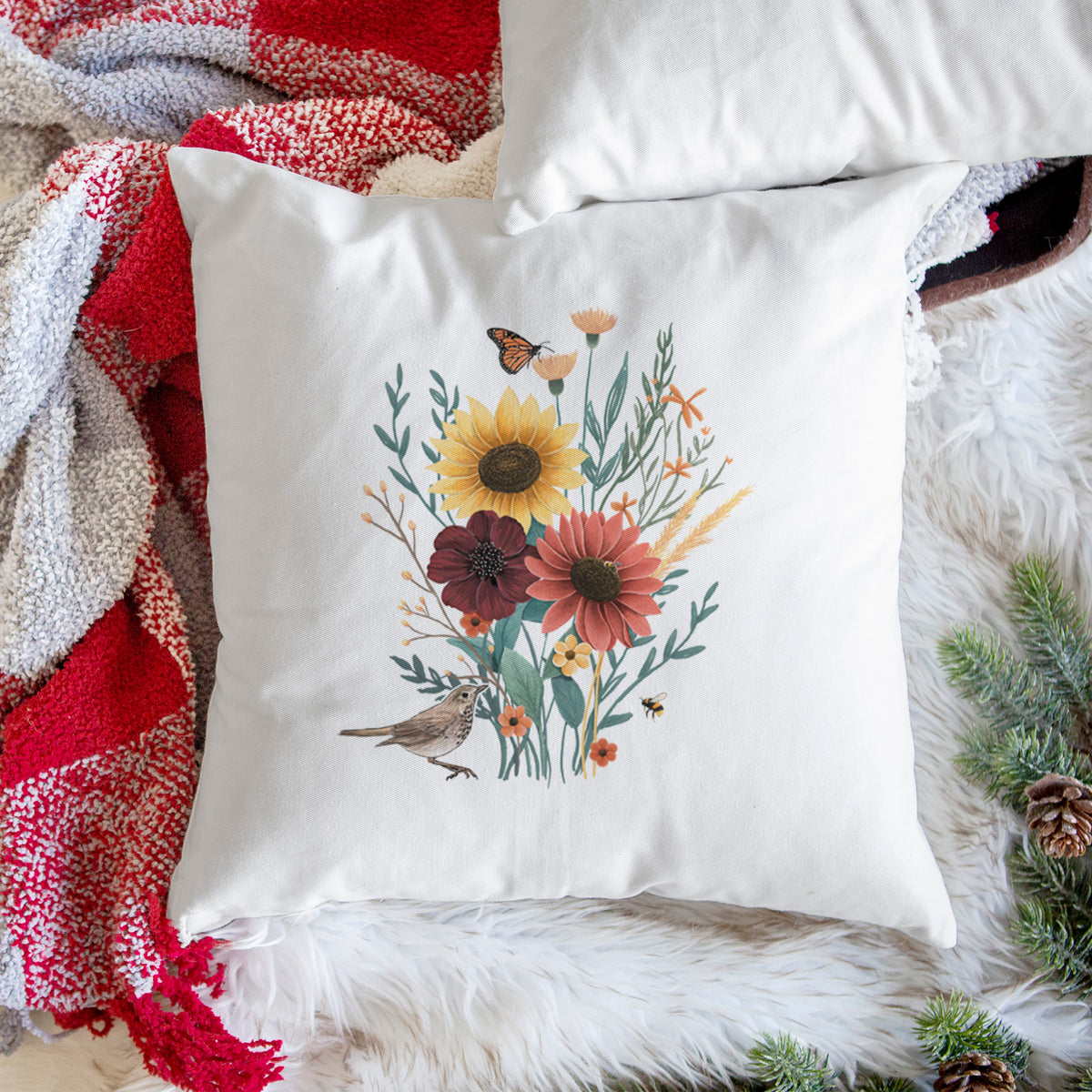 White pillow cover printed with a full-color bouquet of sunflowers, red flowers and wildflowers with butterflies and a small bird, on a woven chair.