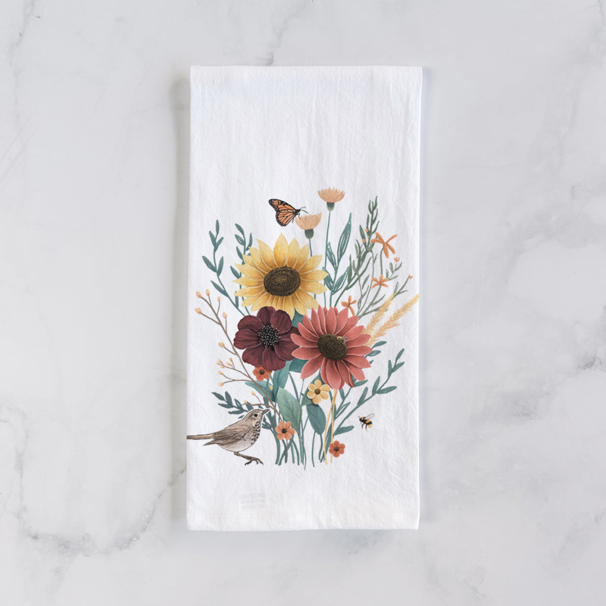 White cotton tea towel laid flat on marble, printed with a full-color bouquet of sunflowers, red flowers and wildflowers with butterflies and a small bird.
