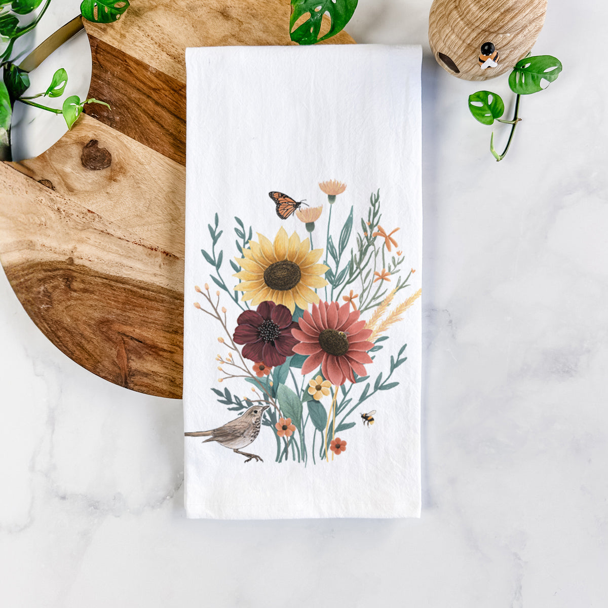White cotton tea towel printed with a full-color bouquet of sunflowers, red flowers and wildflowers with butterflies and a small bird, on a marble counter beside a round wooden cutting board and greenery.
