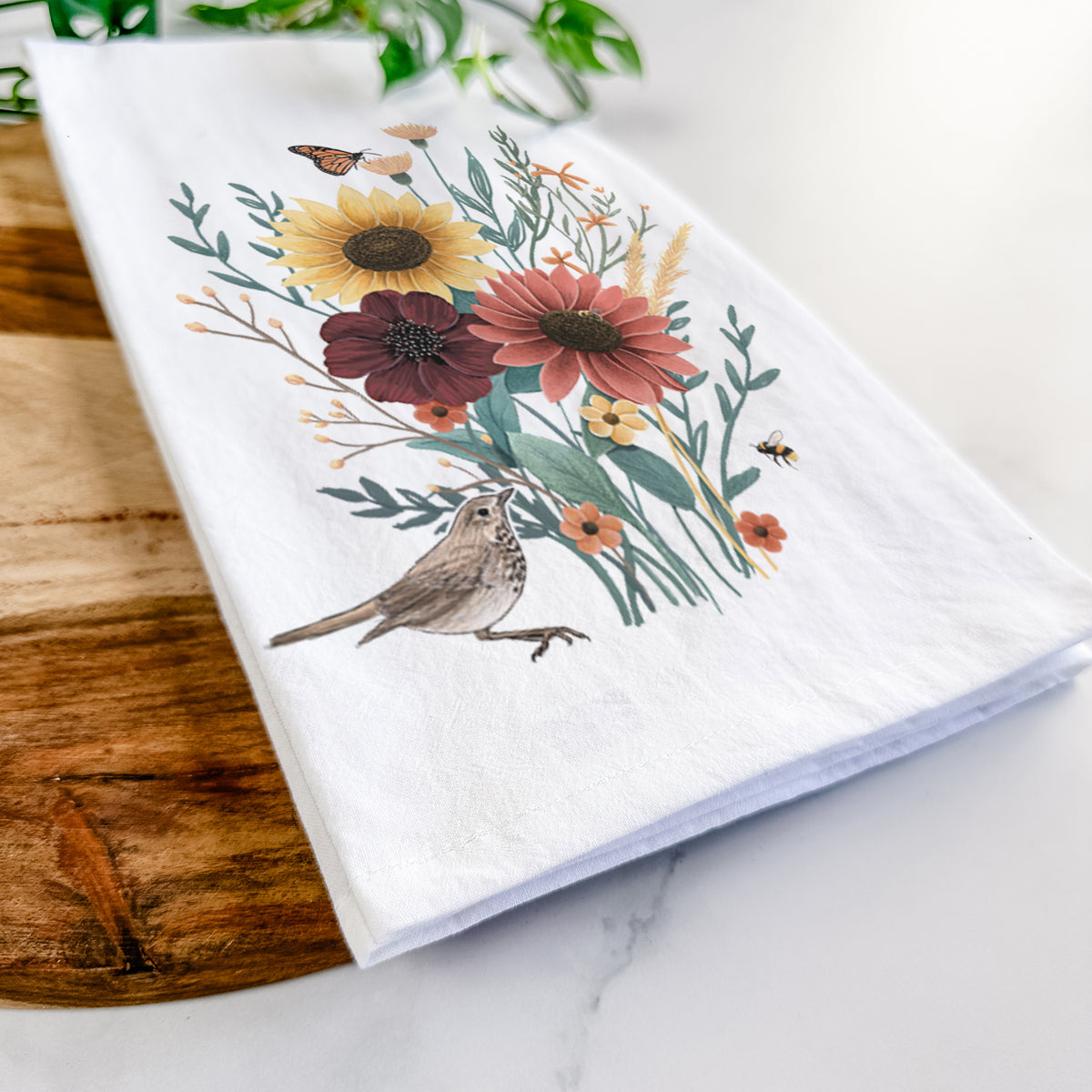 Close-up of a folded white tea towel on a wooden board, showing a full-color bouquet of sunflowers, red flowers and wildflowers with butterflies and a small bird.