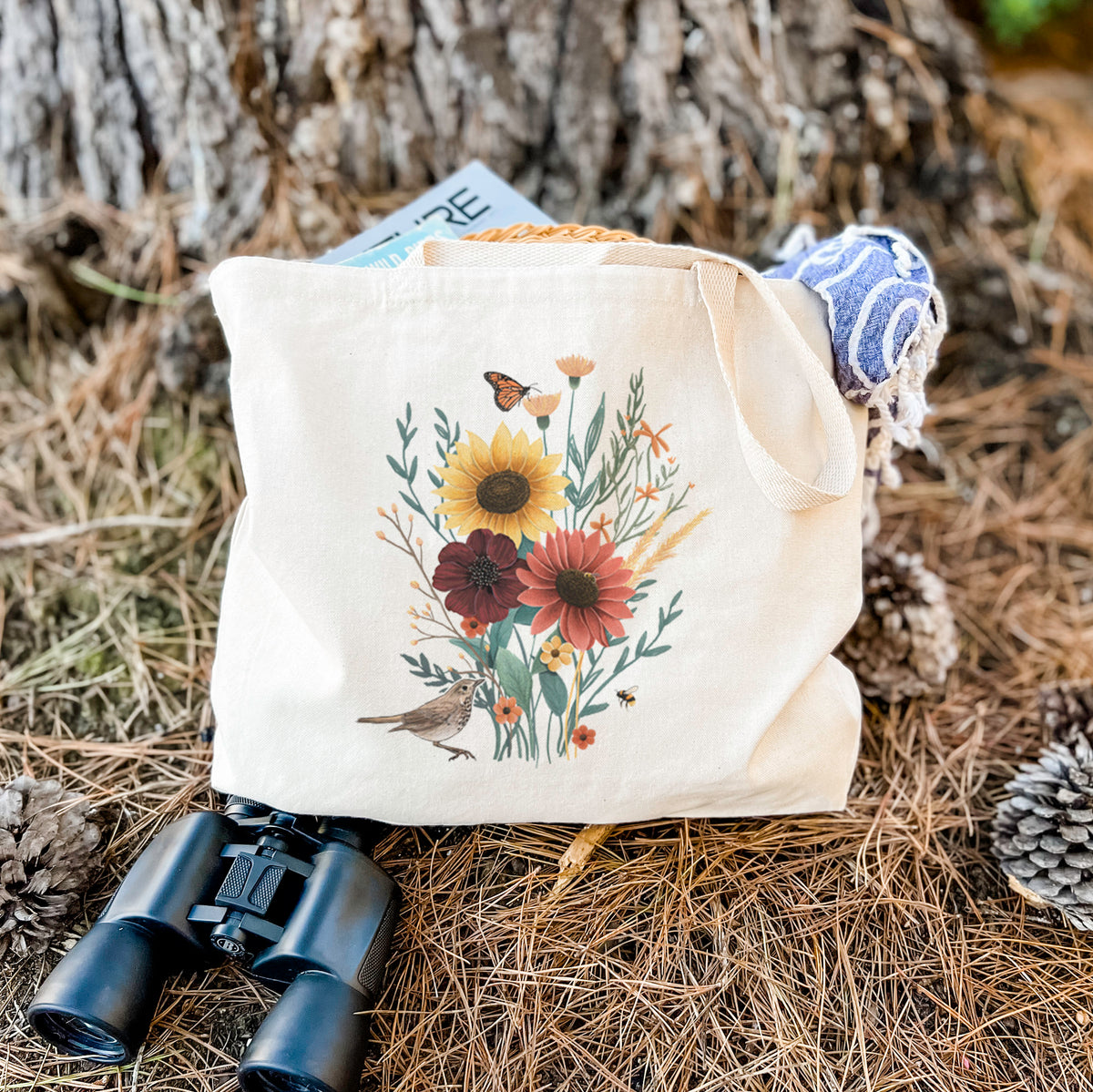 Canvas tote bag printed with a full-color bouquet of sunflowers, red flowers and wildflowers with butterflies and a small bird, resting on dry grass beside a pair of binoculars.