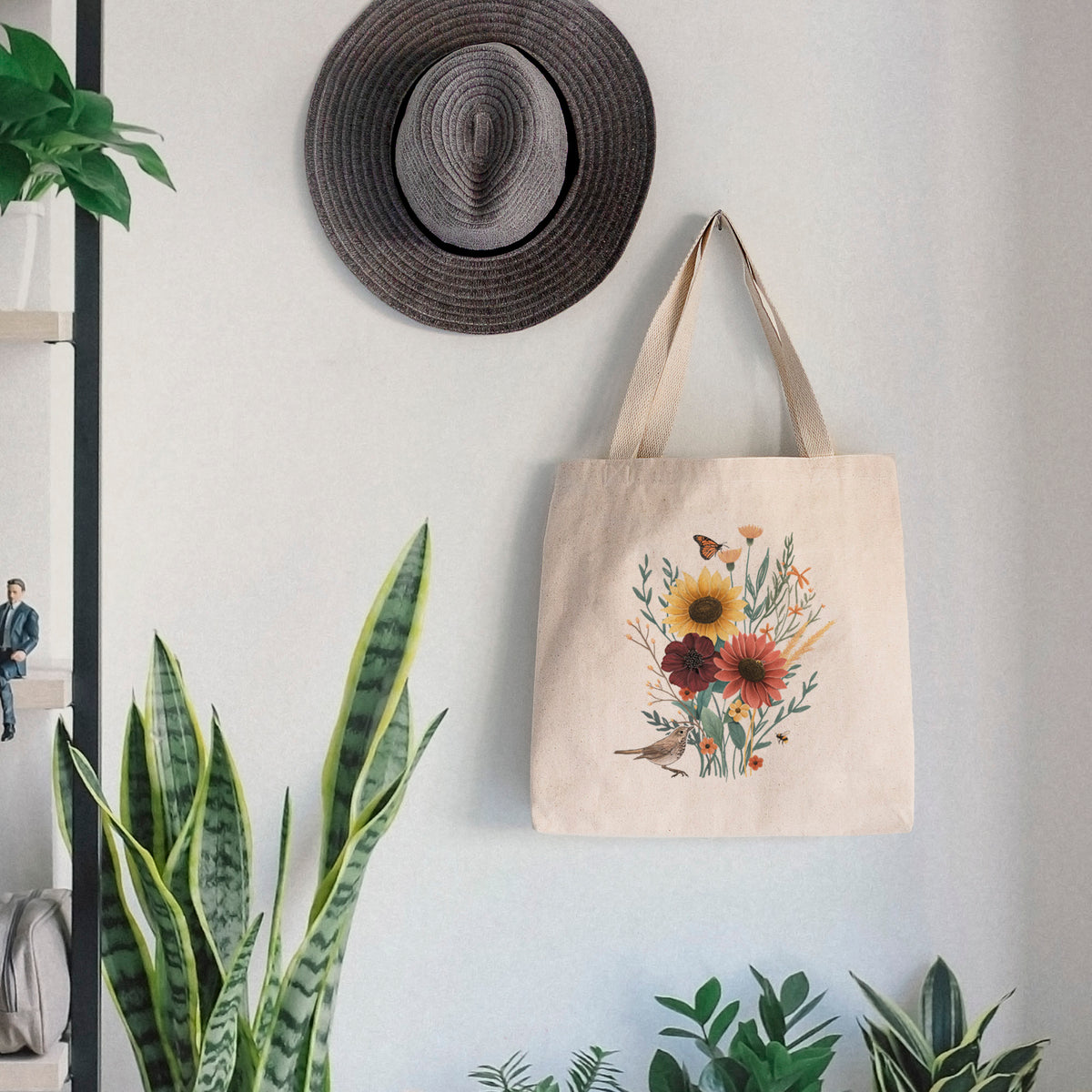 Canvas tote bag printed with a full-color bouquet of sunflowers, red flowers and wildflowers with butterflies and a small bird, hanging on a wall hook below a hat, next to potted plants.