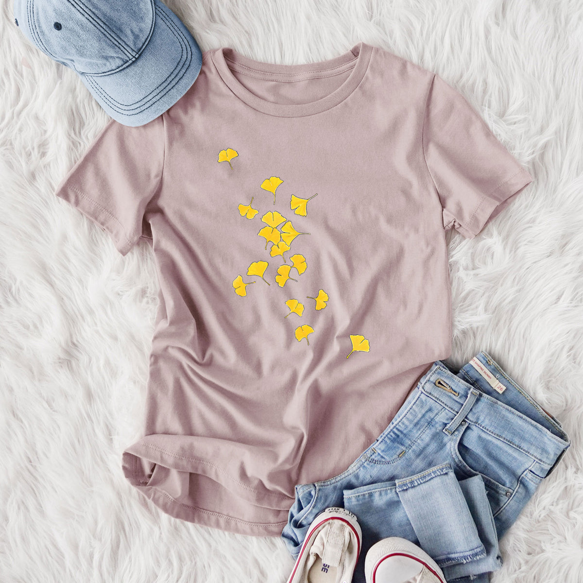 Women's relaxed fit cotton t-shirt printed with a scatter of bright yellow fan-shaped ginkgo leaves falling, styled flat on a white faux fur rug with jeans.