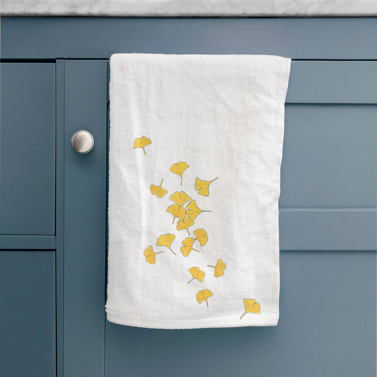 White hand towel printed with a scatter of bright yellow fan-shaped ginkgo leaves falling, hanging on a cabinet door.