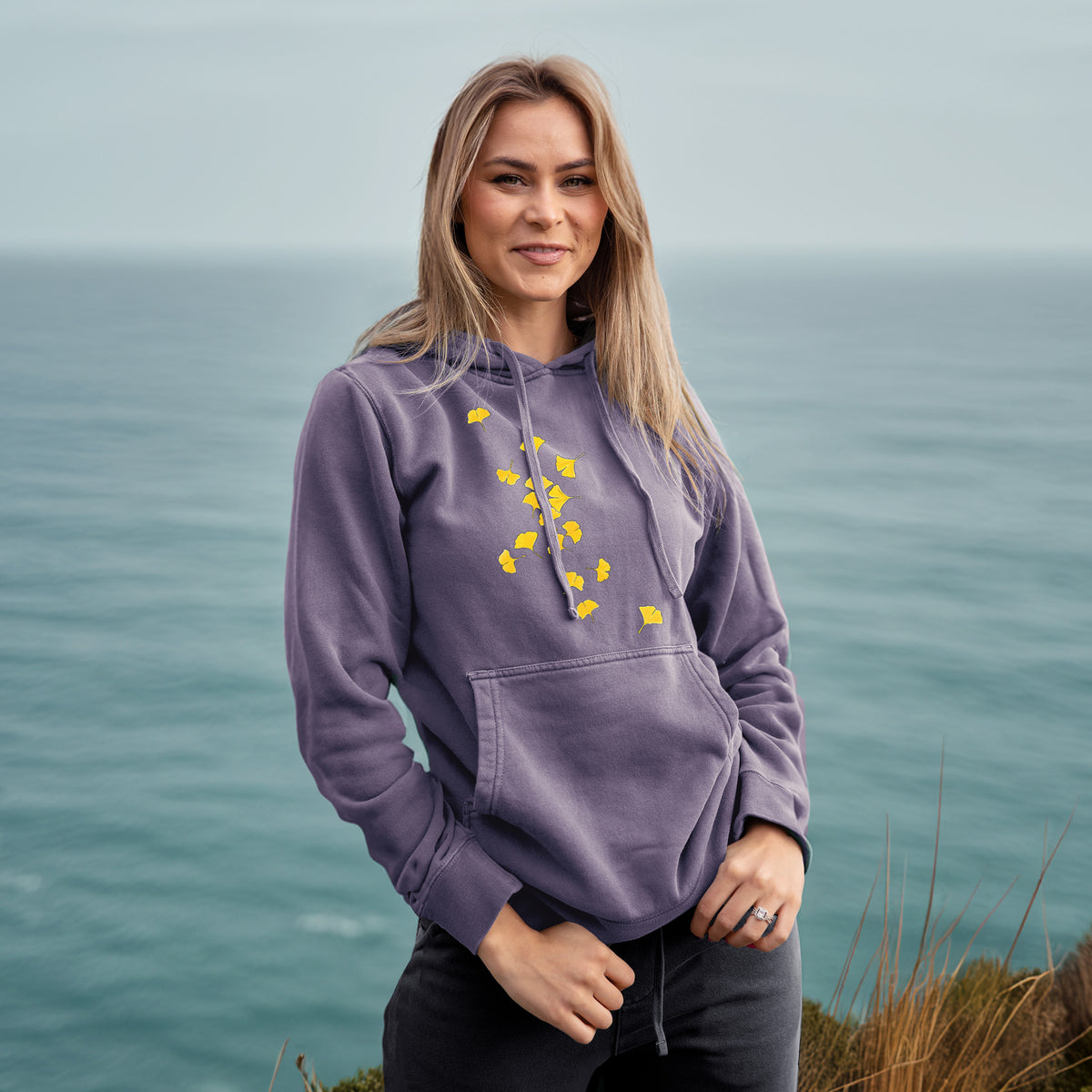 Woman wearing a plum purple pigment dyed hoodie printed with a scatter of bright yellow fan-shaped ginkgo leaves falling.