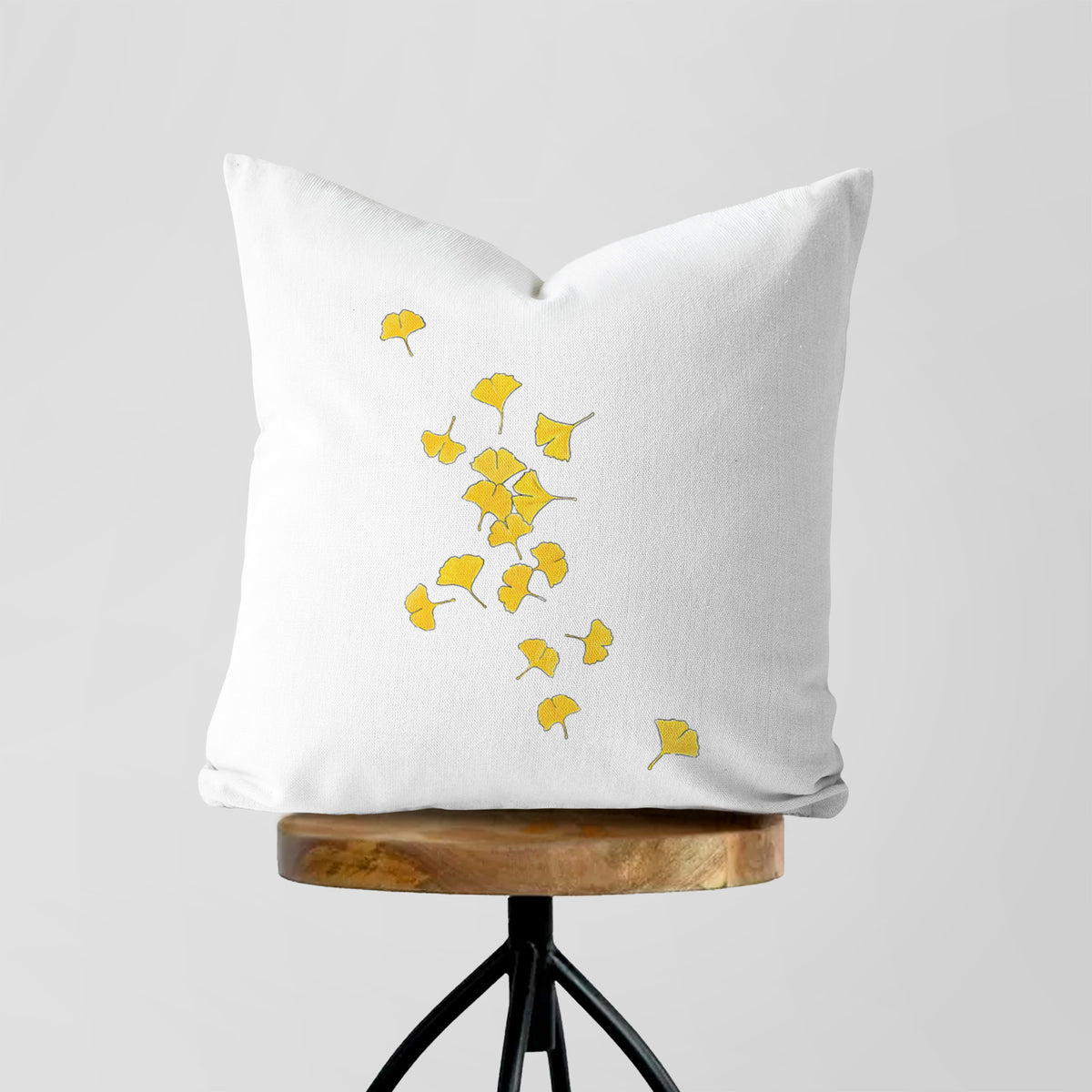 White pillow cover printed with a scatter of bright yellow fan-shaped ginkgo leaves falling, on a wooden stool.