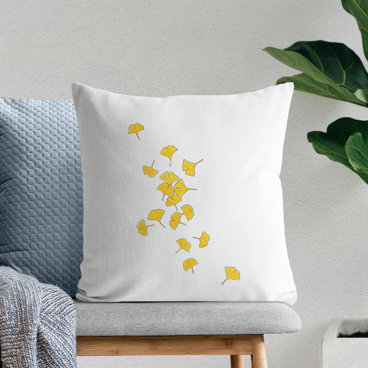 White pillow cover printed with a scatter of bright yellow fan-shaped ginkgo leaves falling, on a bench beside plants.