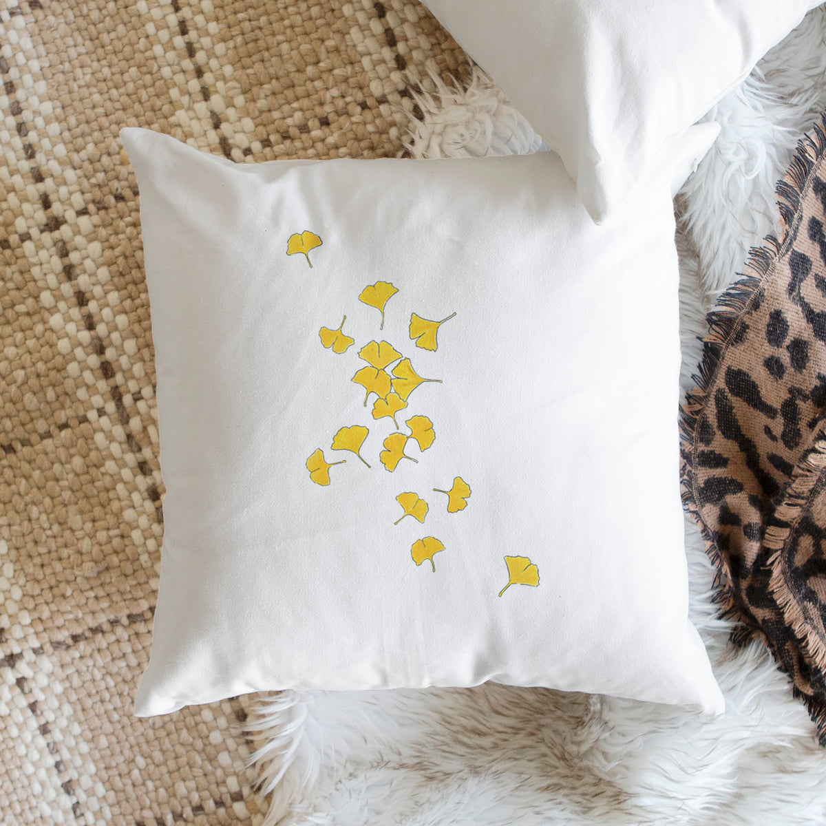White pillow cover printed with a scatter of bright yellow fan-shaped ginkgo leaves falling, on a woven chair.