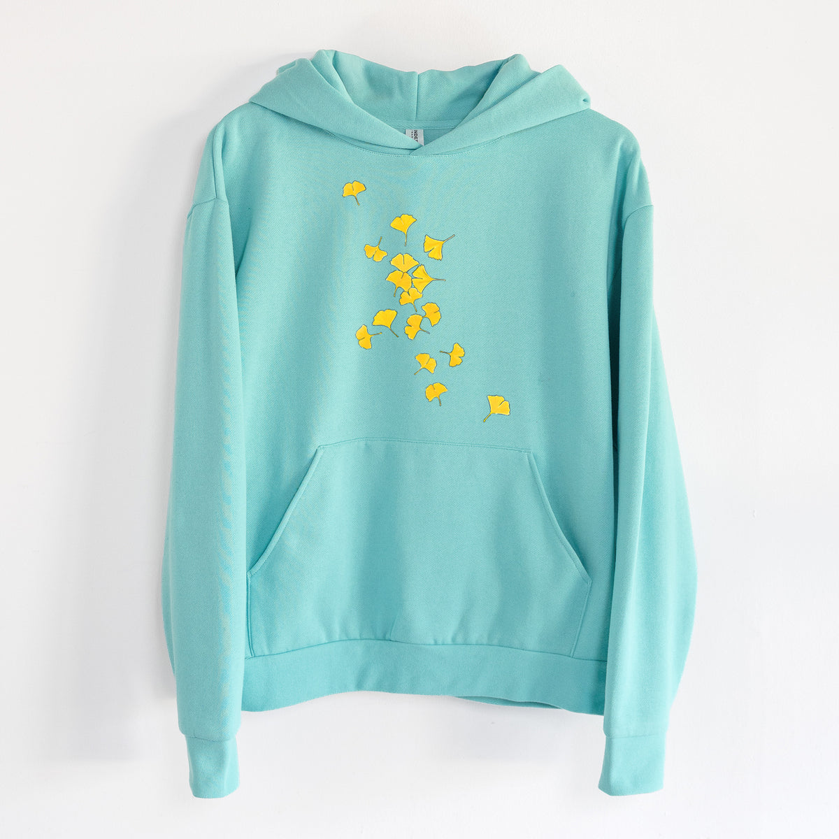 Falling Ginkgo Leaves - Statement Midweight Hoodie in Seafoam, hanging on a white wall, with a front kangaroo pocket and no drawcords.