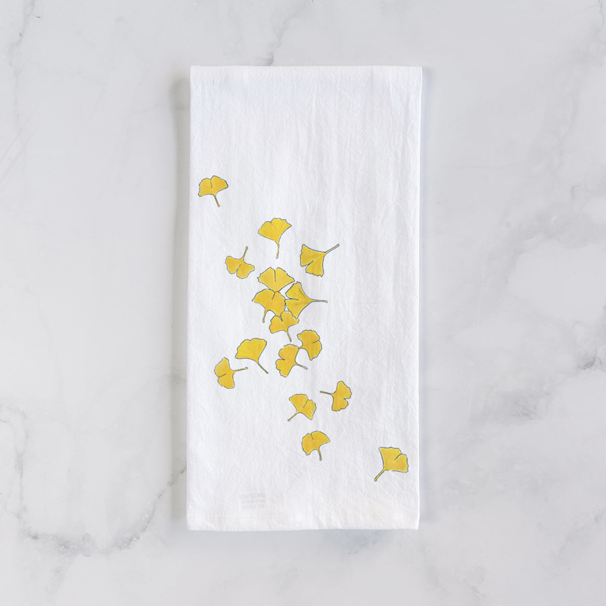 White cotton tea towel laid flat on marble, printed with a scatter of bright yellow fan-shaped ginkgo leaves falling.