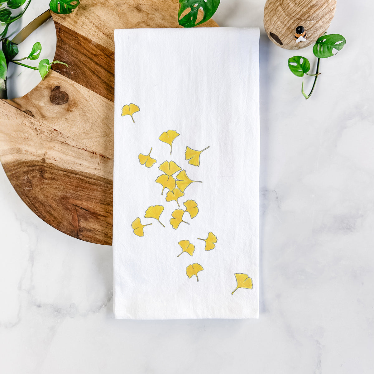 White cotton tea towel printed with a scatter of bright yellow fan-shaped ginkgo leaves falling, on a marble counter beside a round wooden cutting board and greenery.