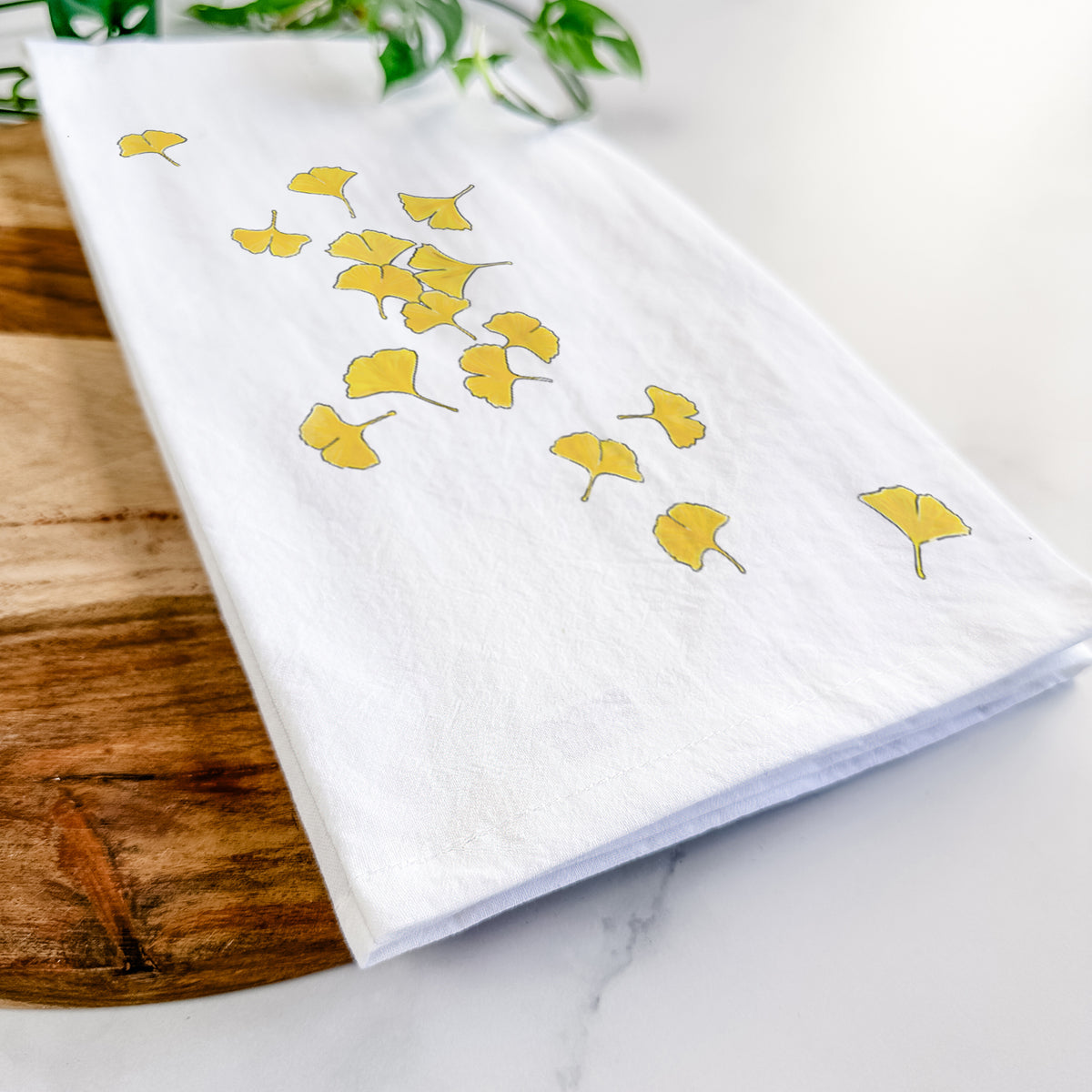 Close-up of a folded white tea towel on a wooden board, showing a scatter of bright yellow fan-shaped ginkgo leaves falling.