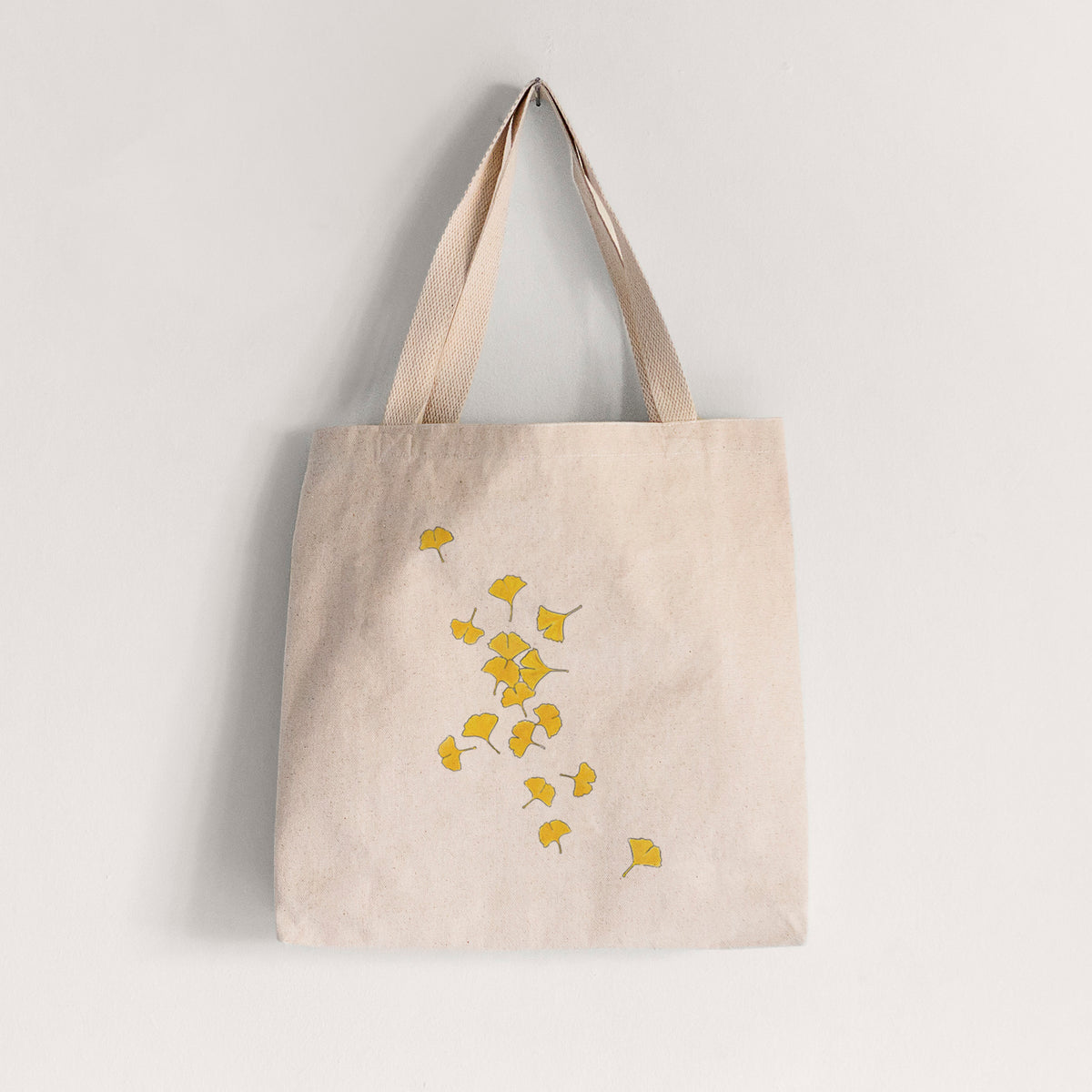 Natural canvas tote bag printed with a scatter of bright yellow fan-shaped ginkgo leaves falling.