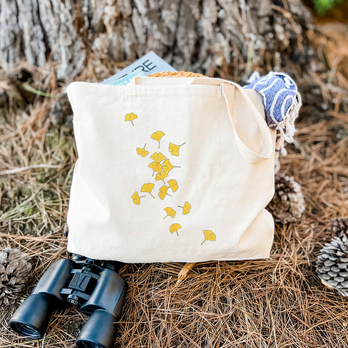 Canvas tote bag printed with a scatter of bright yellow fan-shaped ginkgo leaves falling, resting on dry grass beside a pair of binoculars.