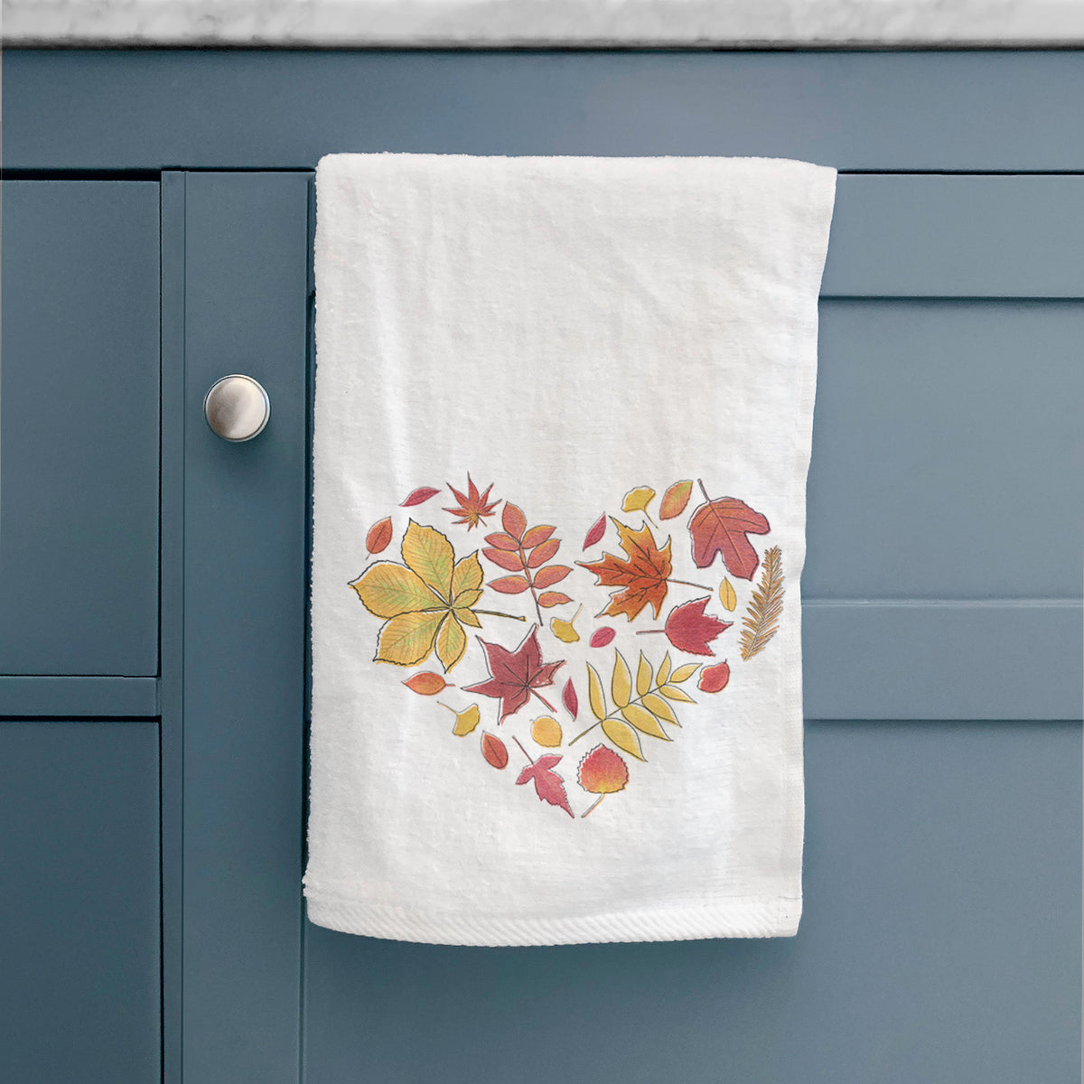 White hand towel printed with a full-color heart shape made of autumn leaves in orange, red and yellow, hanging on a cabinet door.