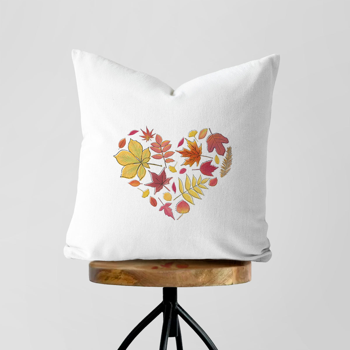 White pillow cover printed with a full-color heart shape made of autumn leaves in orange, red and yellow, on a wooden stool.