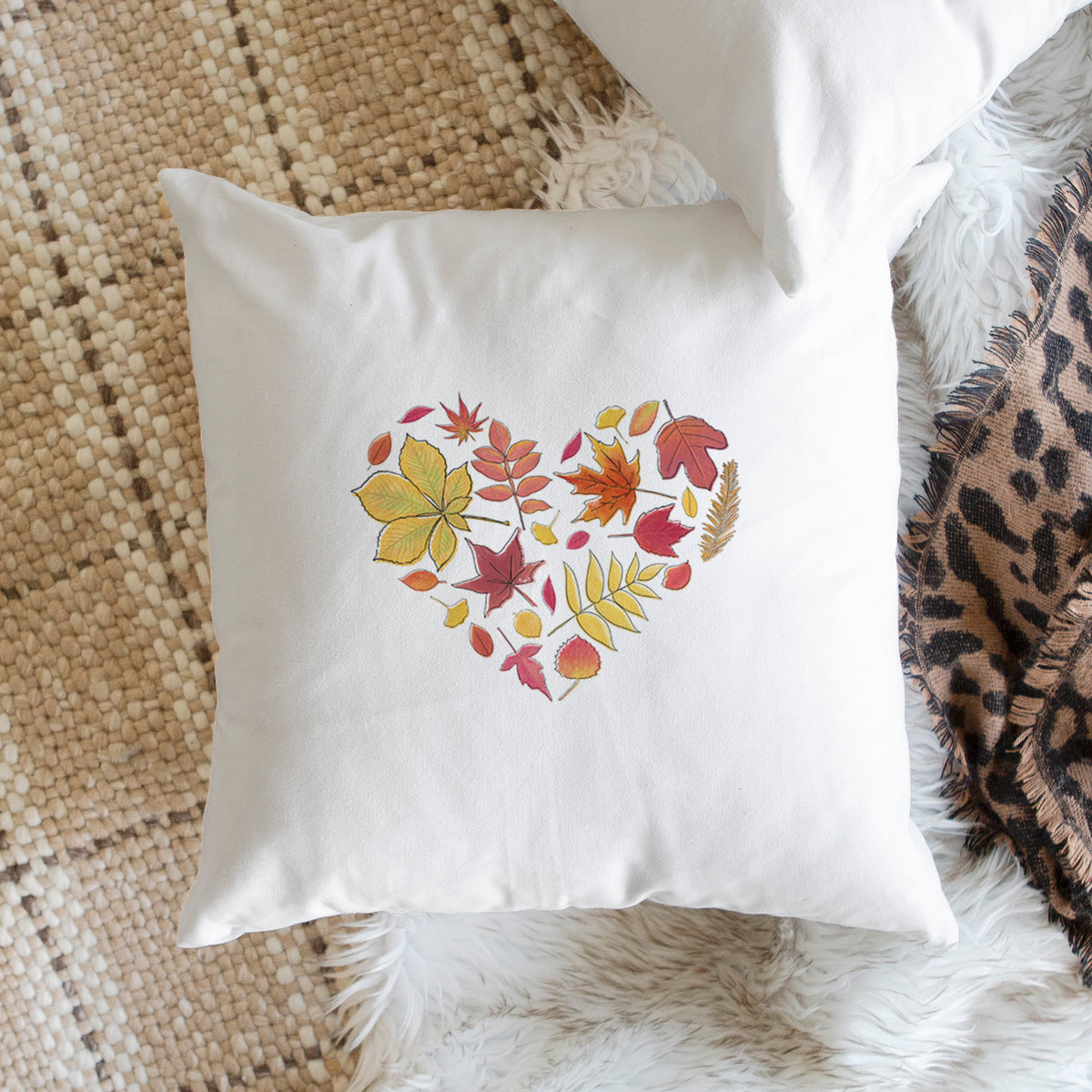 White pillow cover printed with a full-color heart shape made of autumn leaves in orange, red and yellow, on a woven chair.