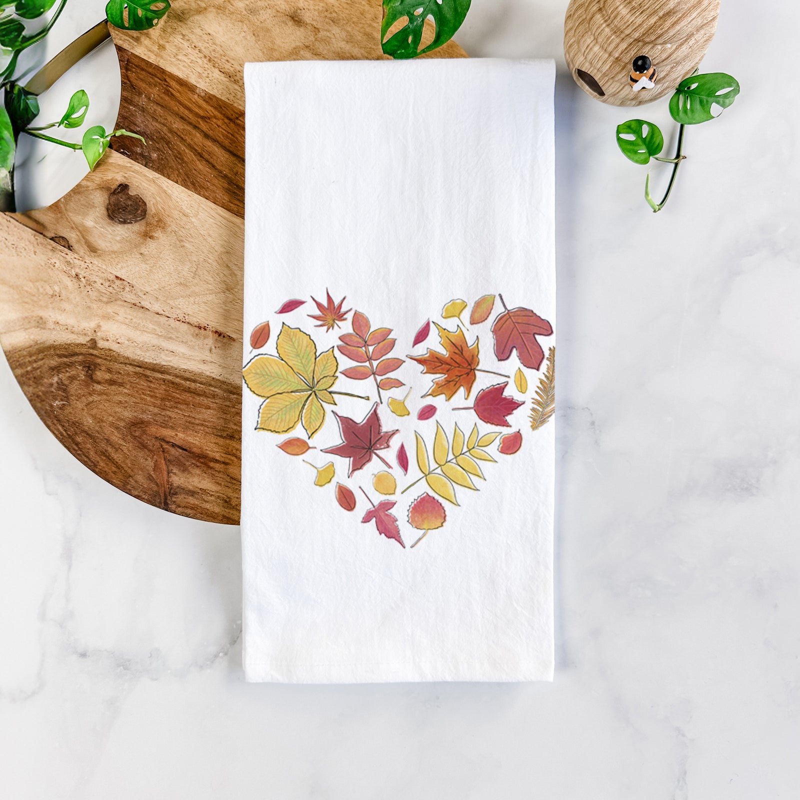 White cotton tea towel printed with a full-color heart shape made of autumn leaves in orange, red and yellow, on a marble counter beside a round wooden cutting board and greenery.