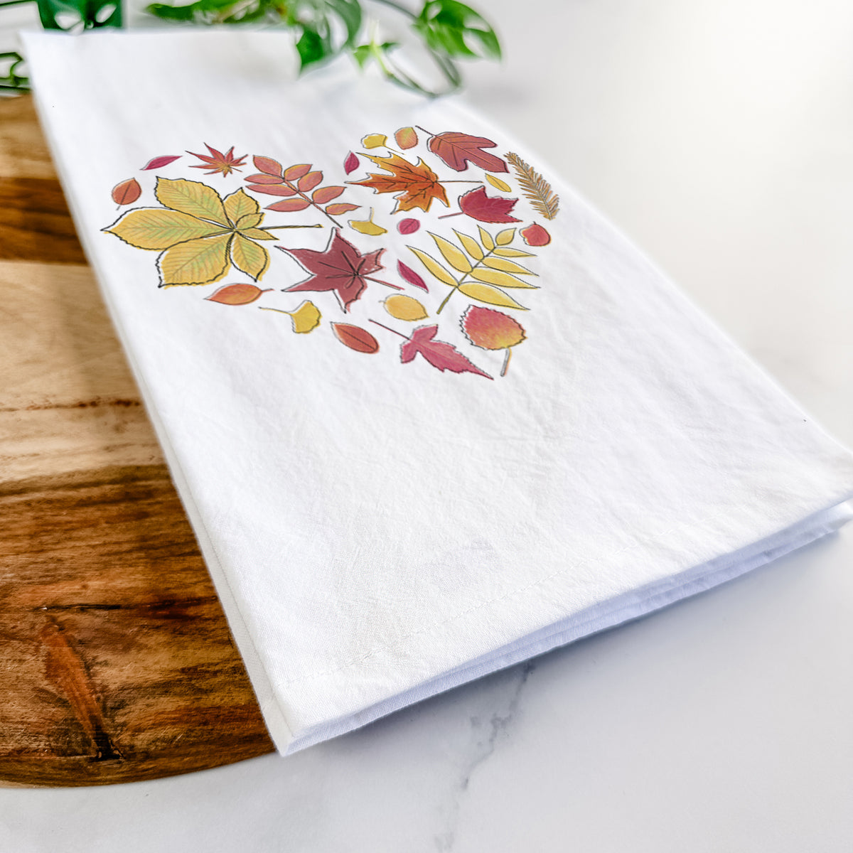 Close-up of a folded white tea towel on a wooden board, showing a full-color heart shape made of autumn leaves in orange, red and yellow.