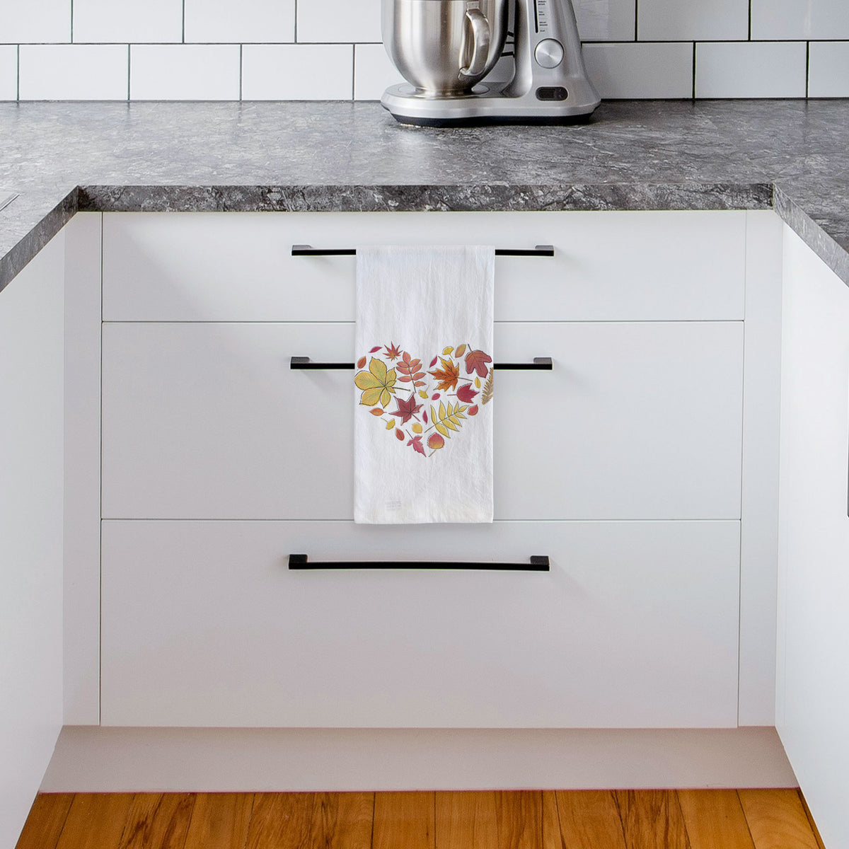 White tea towel printed with a full-color heart shape made of autumn leaves in orange, red and yellow, hanging from a drawer handle in a bright white kitchen.