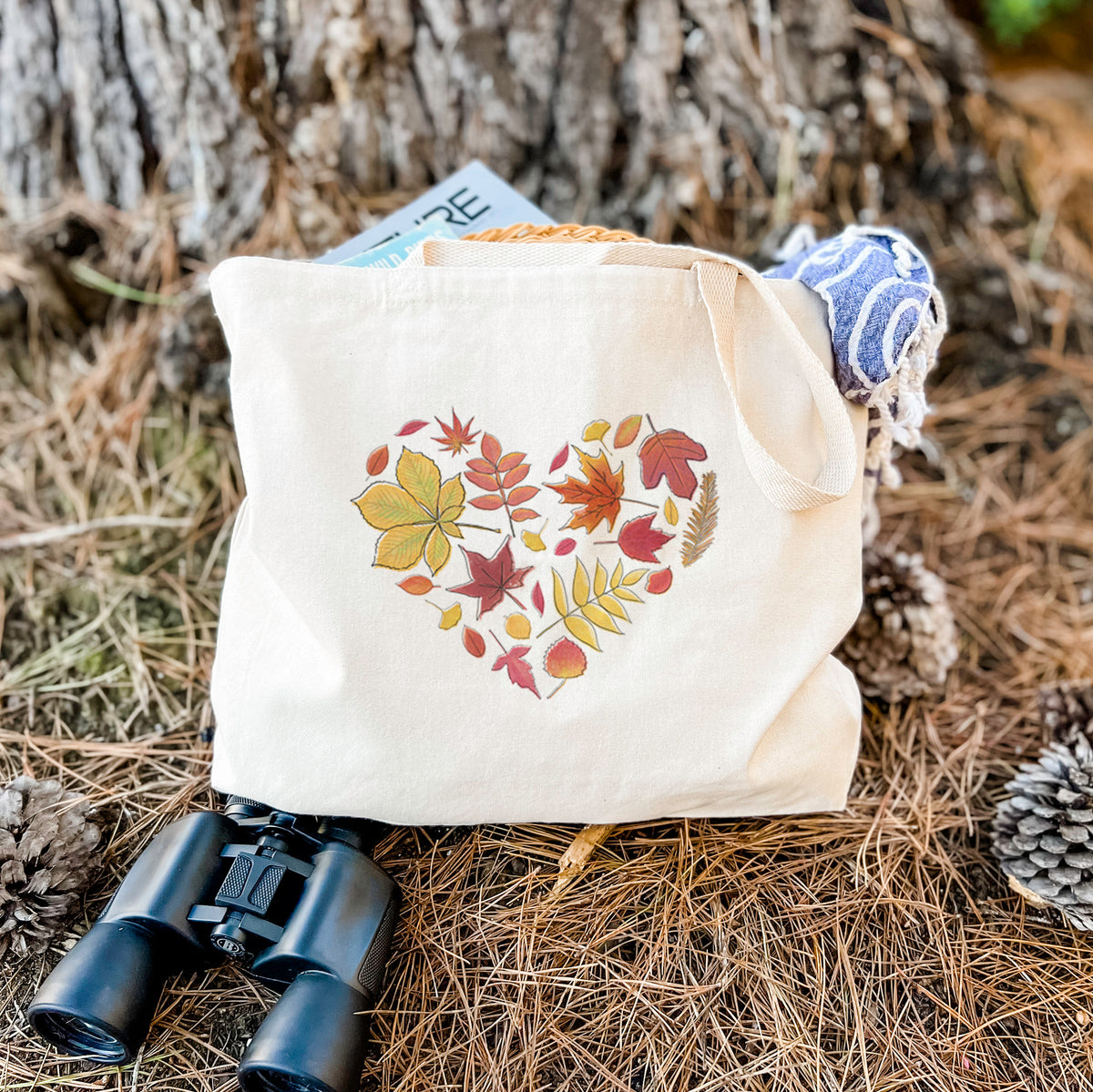 Canvas tote bag printed with a full-color heart shape made of autumn leaves in orange, red and yellow, resting on dry grass beside a pair of binoculars.