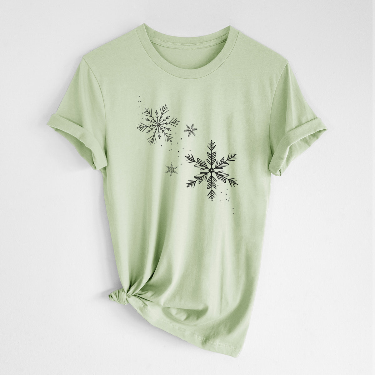 Spring green lightweight cotton t-shirt laid flat with the sleeves rolled, printed with delicate snowflakes of different sizes falling.