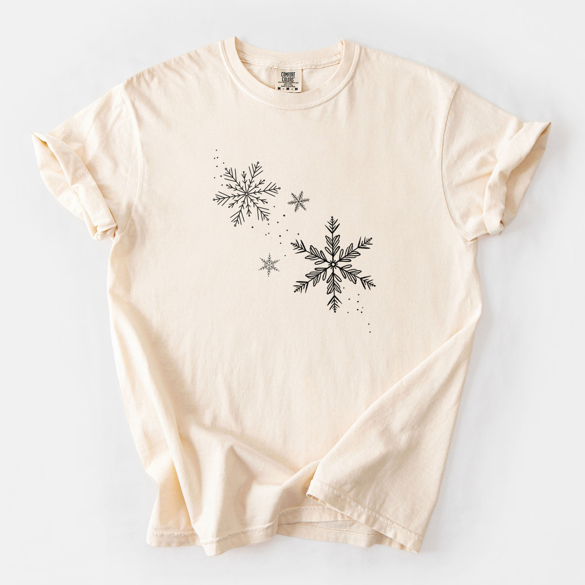 Ivory Comfort Colors t-shirt laid flat, printed with delicate snowflakes of different sizes falling.