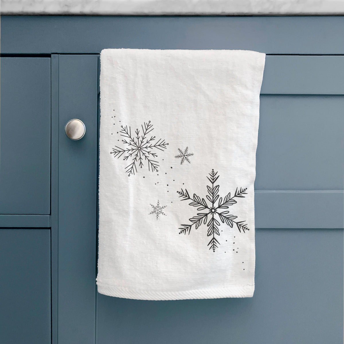 White hand towel printed with delicate snowflakes of different sizes falling, hanging on a cabinet door.