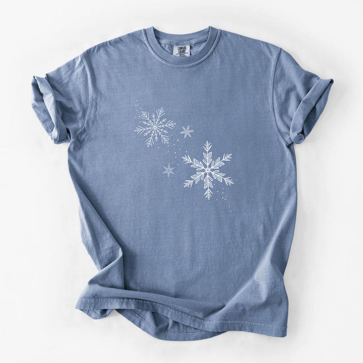 Blue jean Comfort Colors t-shirt laid flat, printed with delicate white snowflakes of different sizes falling.