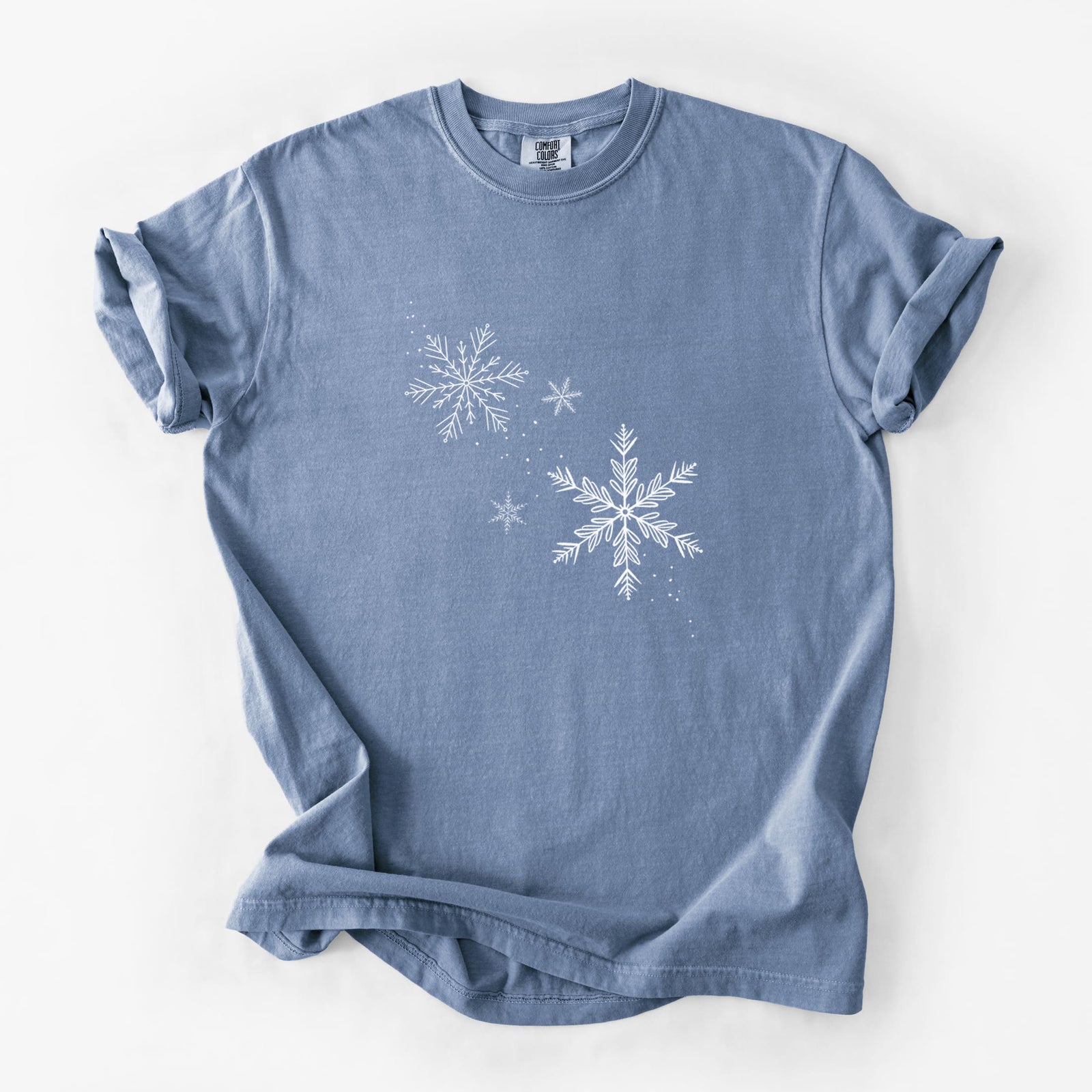 Blue jean Comfort Colors t-shirt laid flat, printed with delicate white snowflakes of different sizes falling.