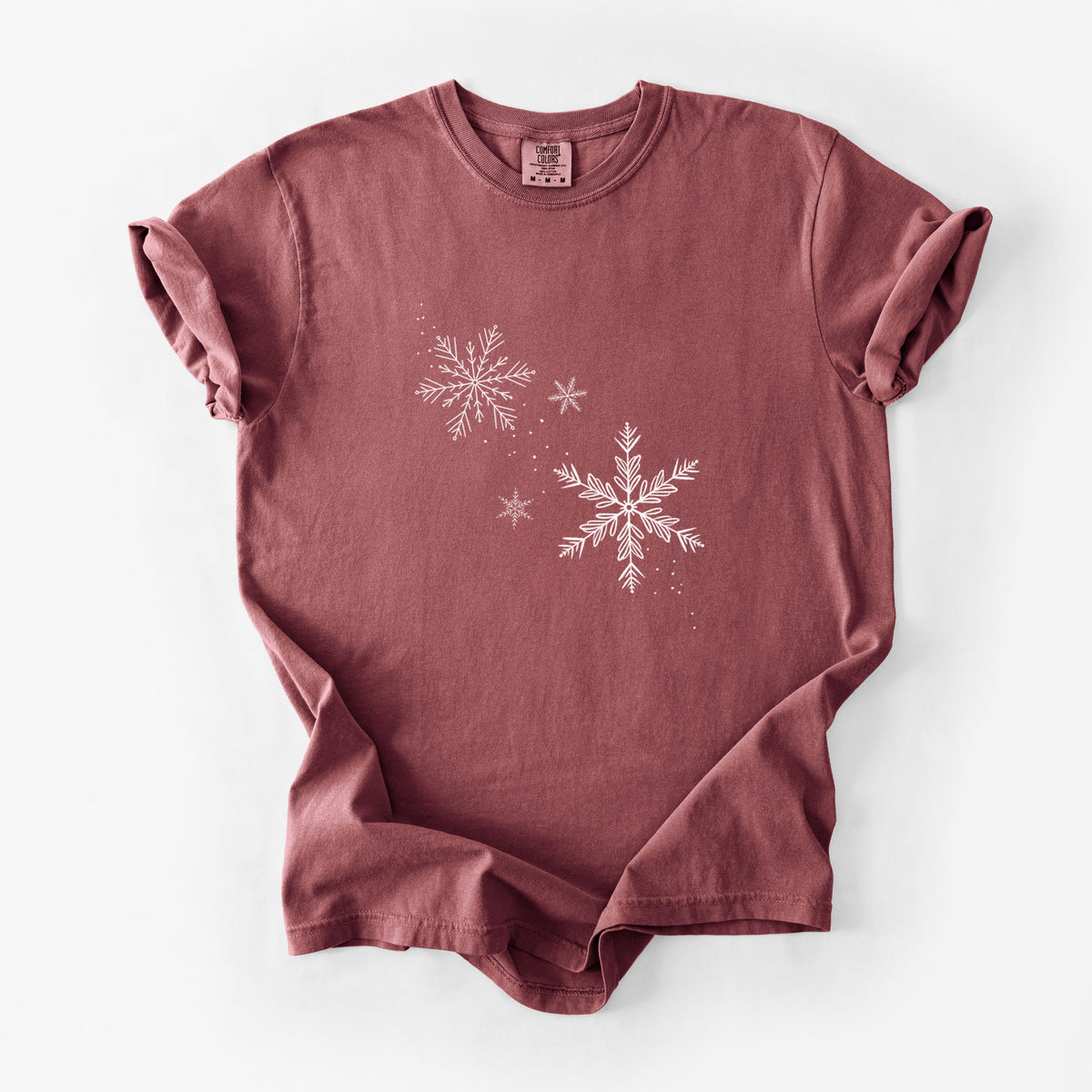 Brick red Comfort Colors t-shirt laid flat, printed with delicate white snowflakes of different sizes falling.