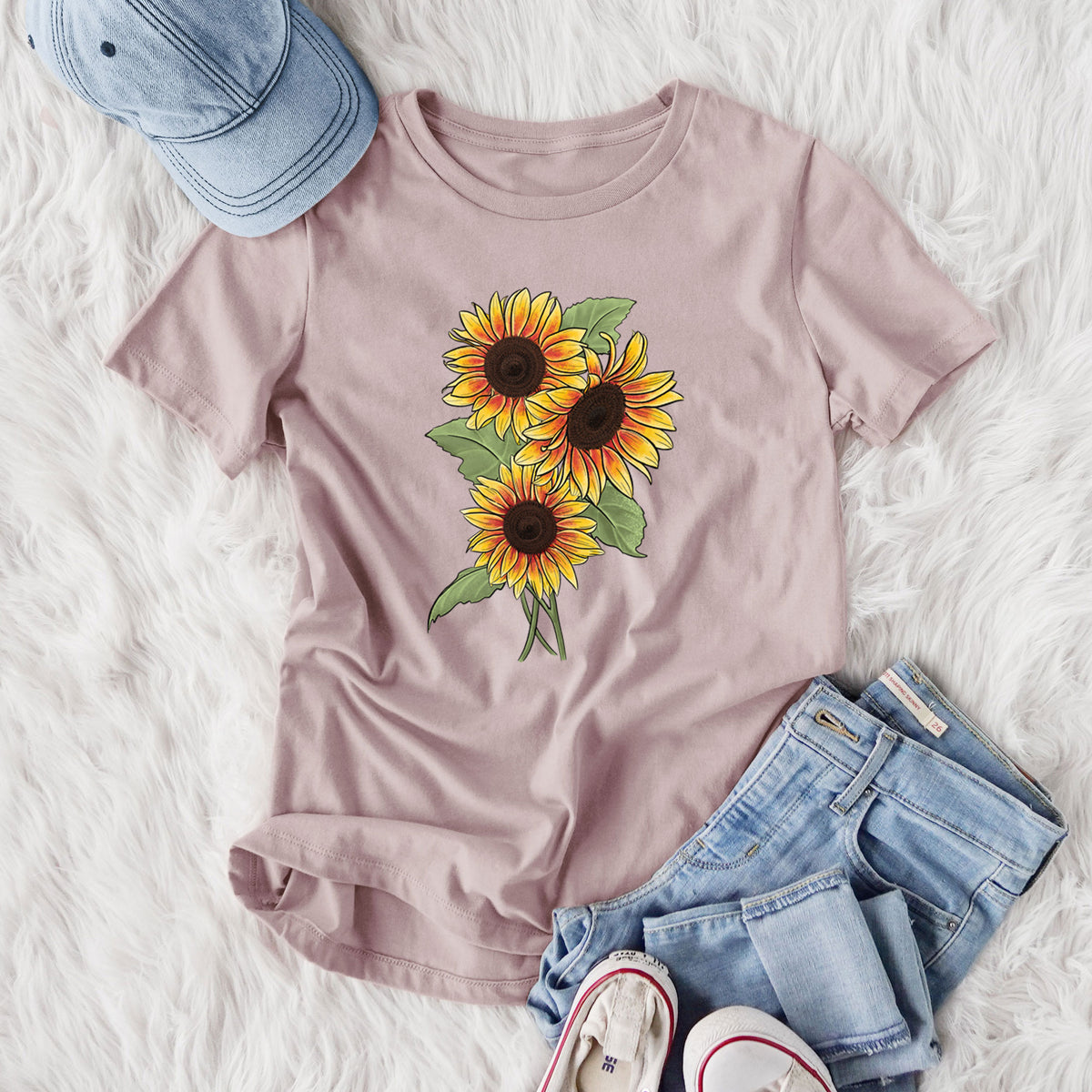 Women's relaxed fit cotton t-shirt printed with a full-color illustration of a bunch of yellow and orange sunflowers, styled flat on a white faux fur rug with jeans.