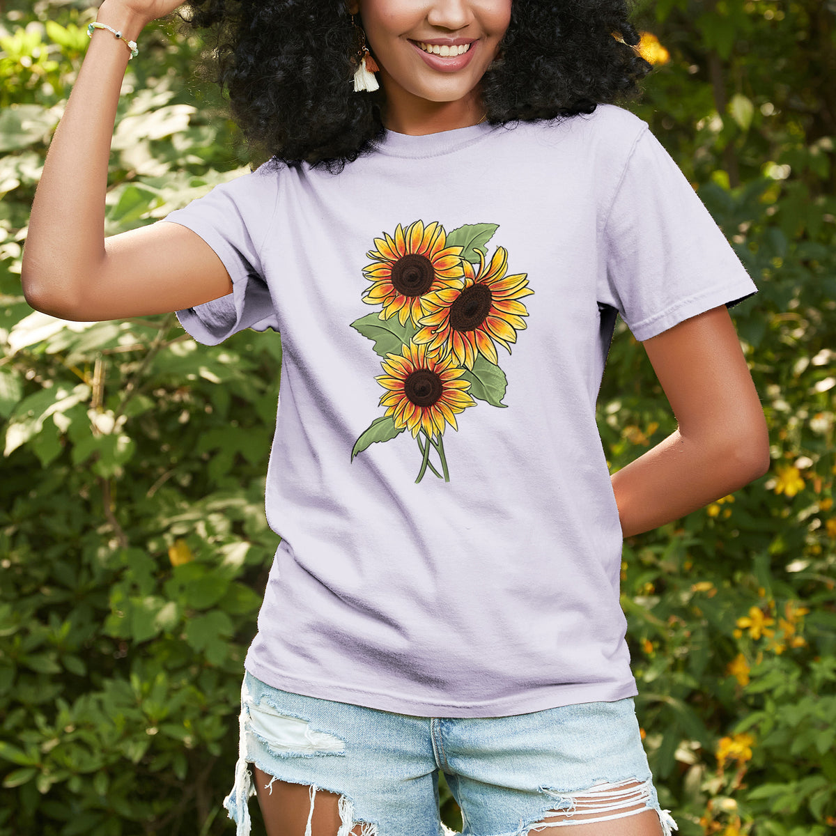 Woman wearing a Comfort Colors t-shirt printed with a full-color illustration of a bunch of yellow and orange sunflowers, outdoors.