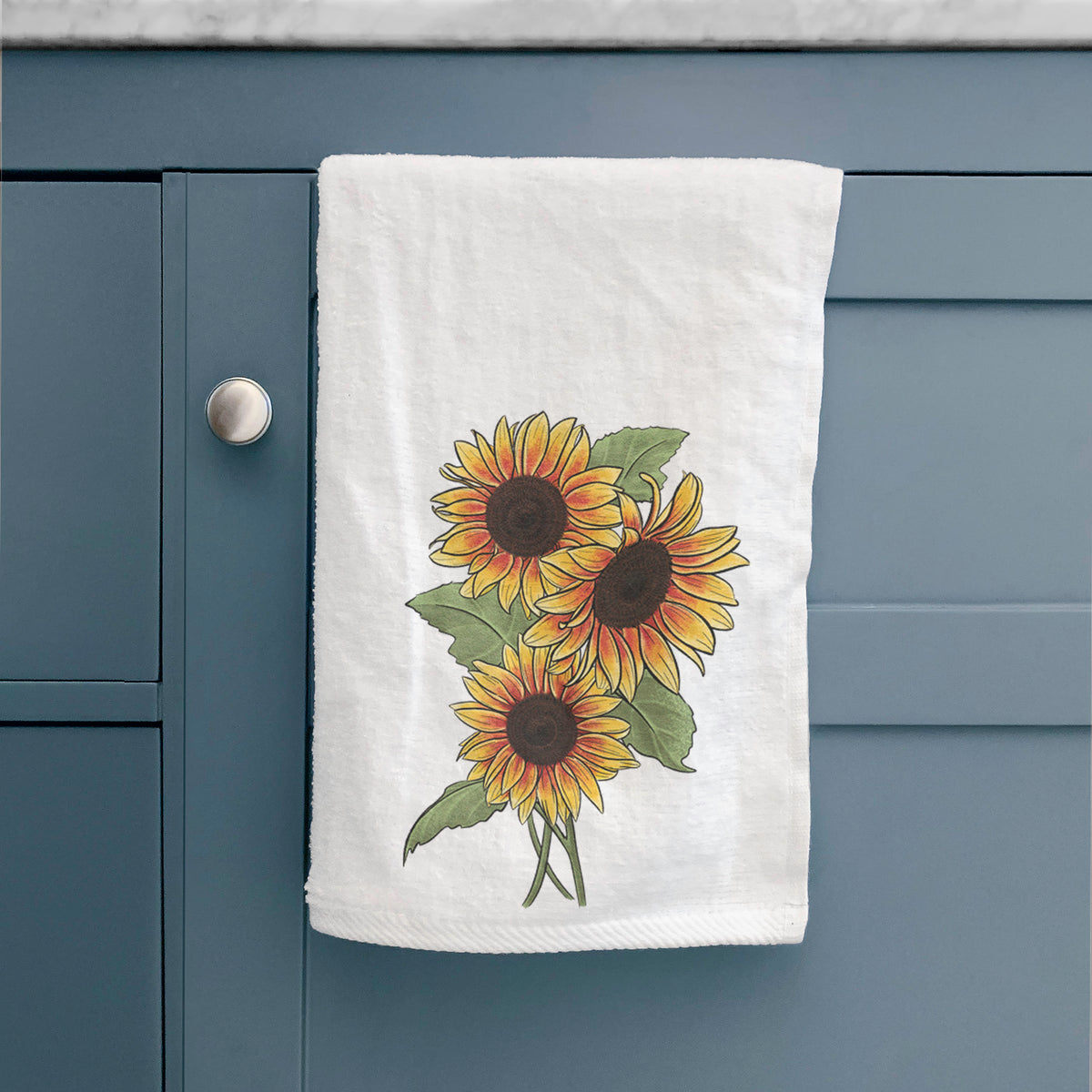 White hand towel printed with a full-color illustration of a bunch of yellow and orange sunflowers, hanging on a cabinet door.