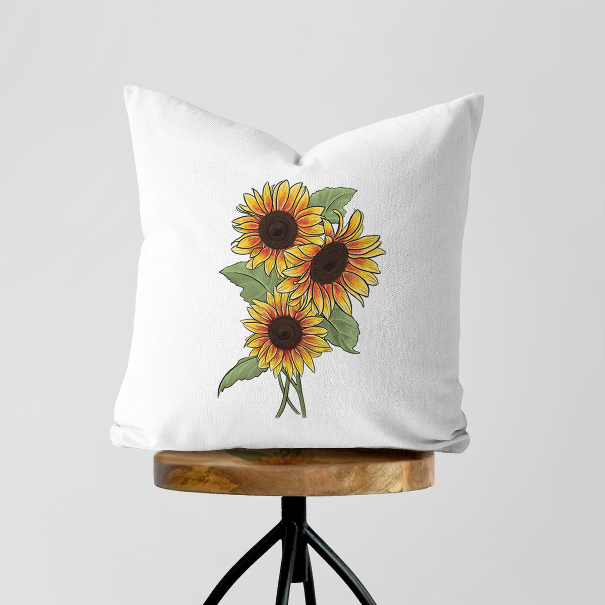 White pillow cover printed with a full-color illustration of a bunch of yellow and orange sunflowers, on a wooden stool.
