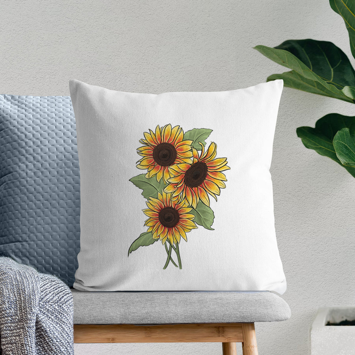 White pillow cover printed with a full-color illustration of a bunch of yellow and orange sunflowers, on a bench beside plants.