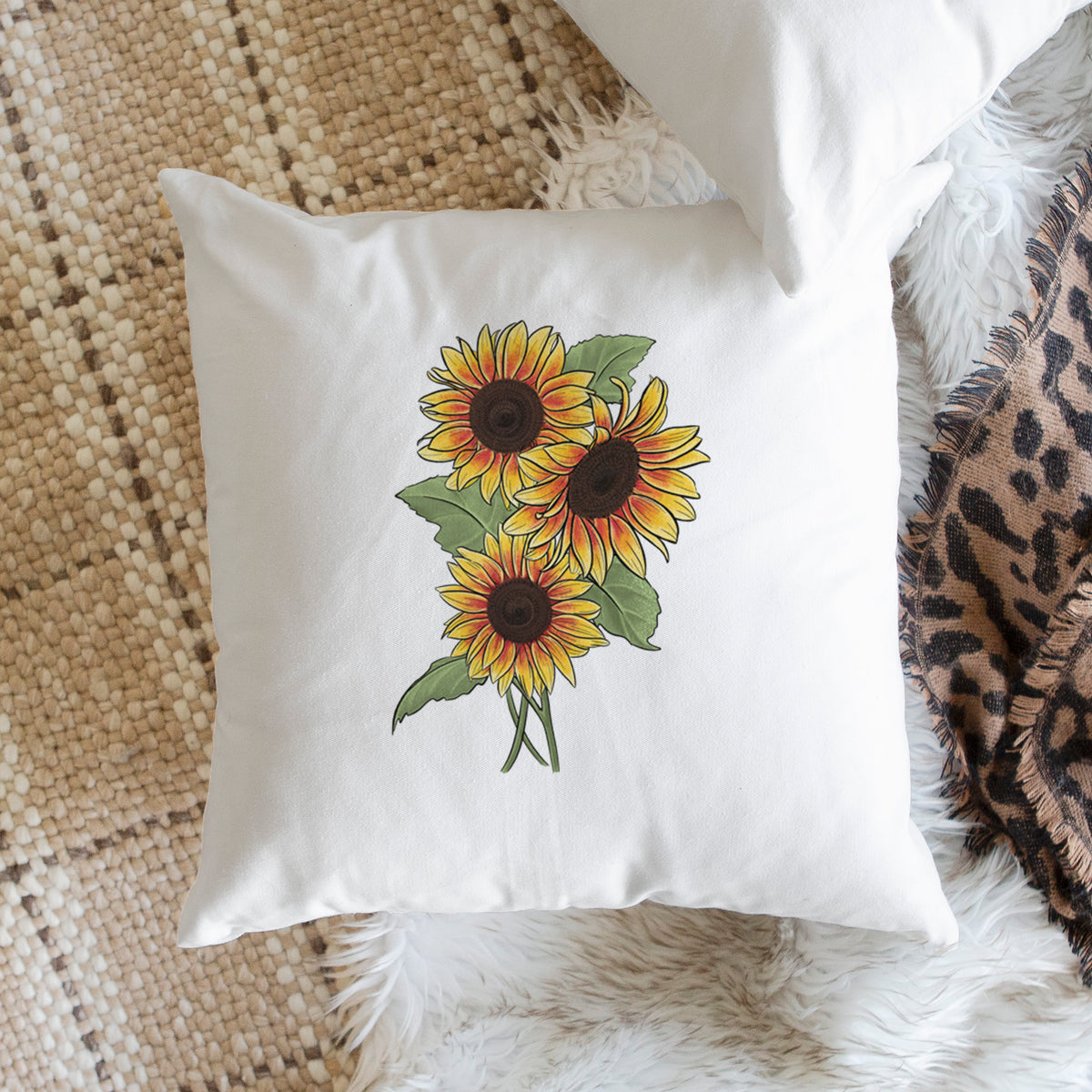 White pillow cover printed with a full-color illustration of a bunch of yellow and orange sunflowers, on a woven chair.