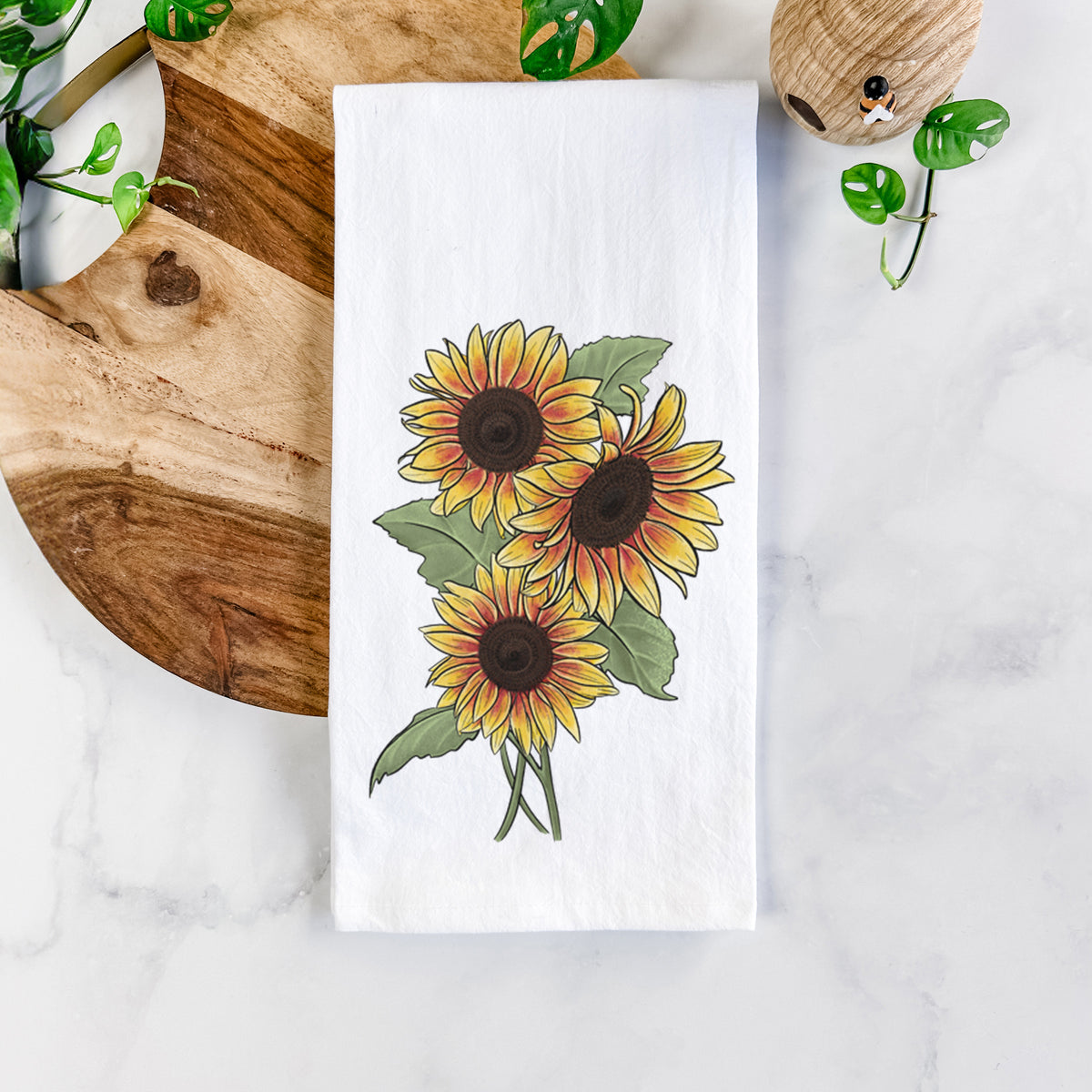 White cotton tea towel printed with a full-color illustration of a bunch of yellow and orange sunflowers, on a marble counter beside a round wooden cutting board and greenery.