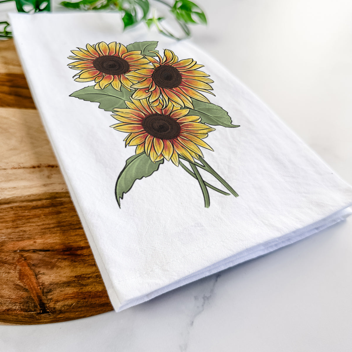 Close-up of a folded white tea towel on a wooden board, showing a full-color illustration of a bunch of yellow and orange sunflowers.